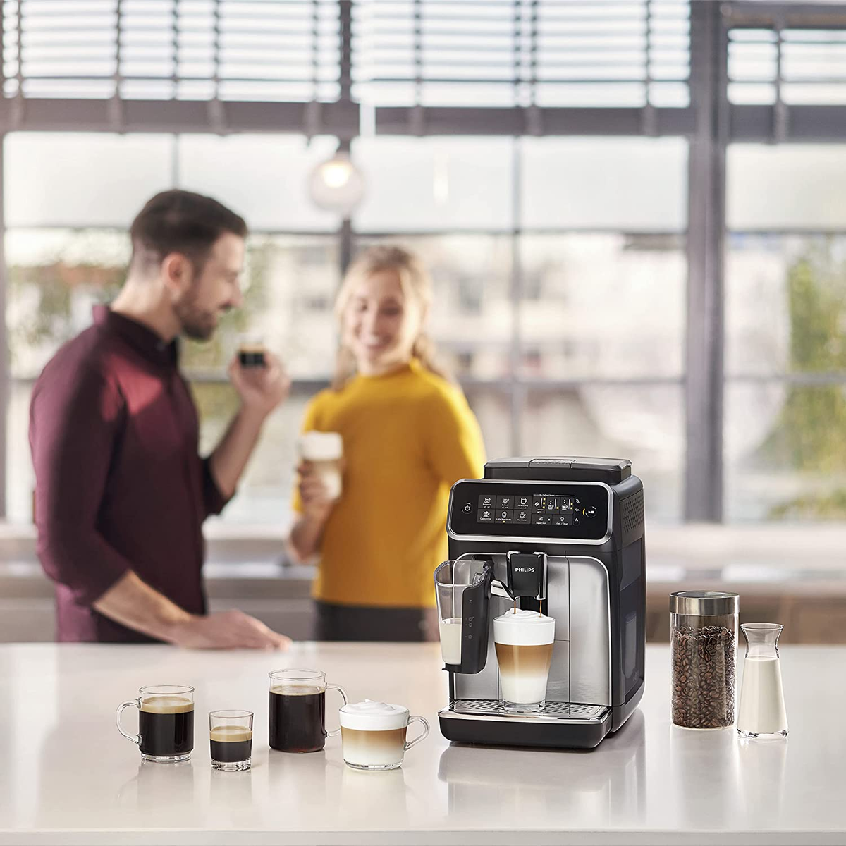 Latte ve fincanlı kahve makinesi. Arka planda iki kişi. Beyaz tezgah.