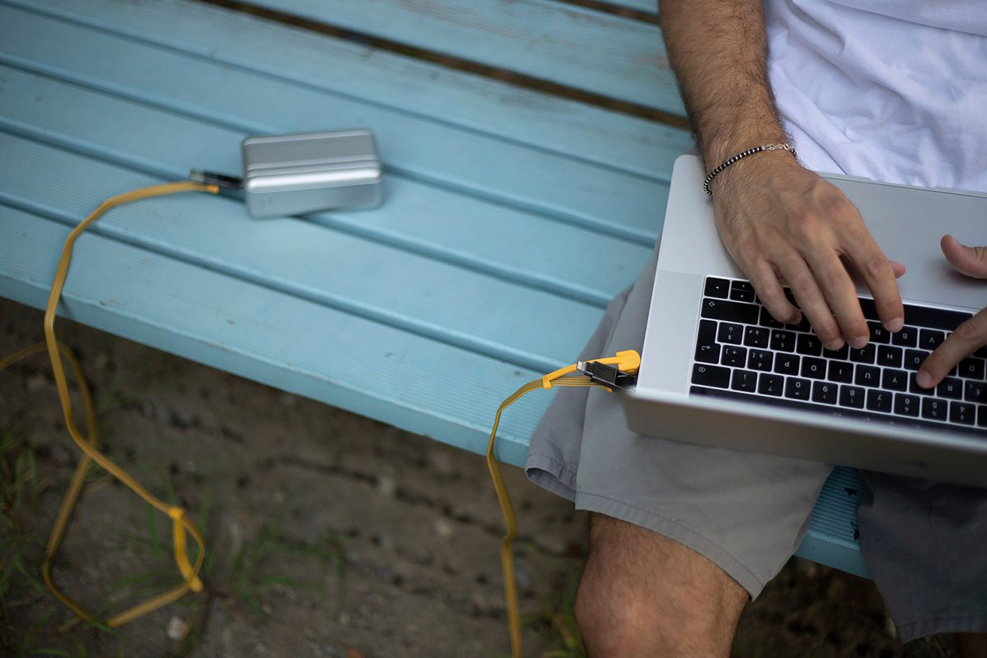 Un homme utilise un ordinateur portable sur un banc bleu. Un appareil argenté et un câble jaune sont à proximité.