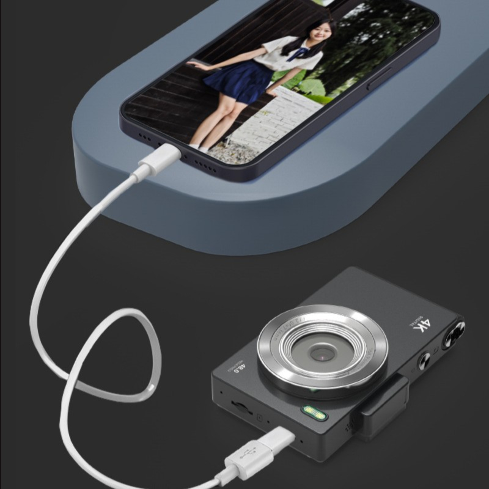 Schwarze Digitalkamera mit weißem Kabel an ein Telefon angeschlossen. Telefon zeigt ein Frauenbild. Grauer Hintergrund.