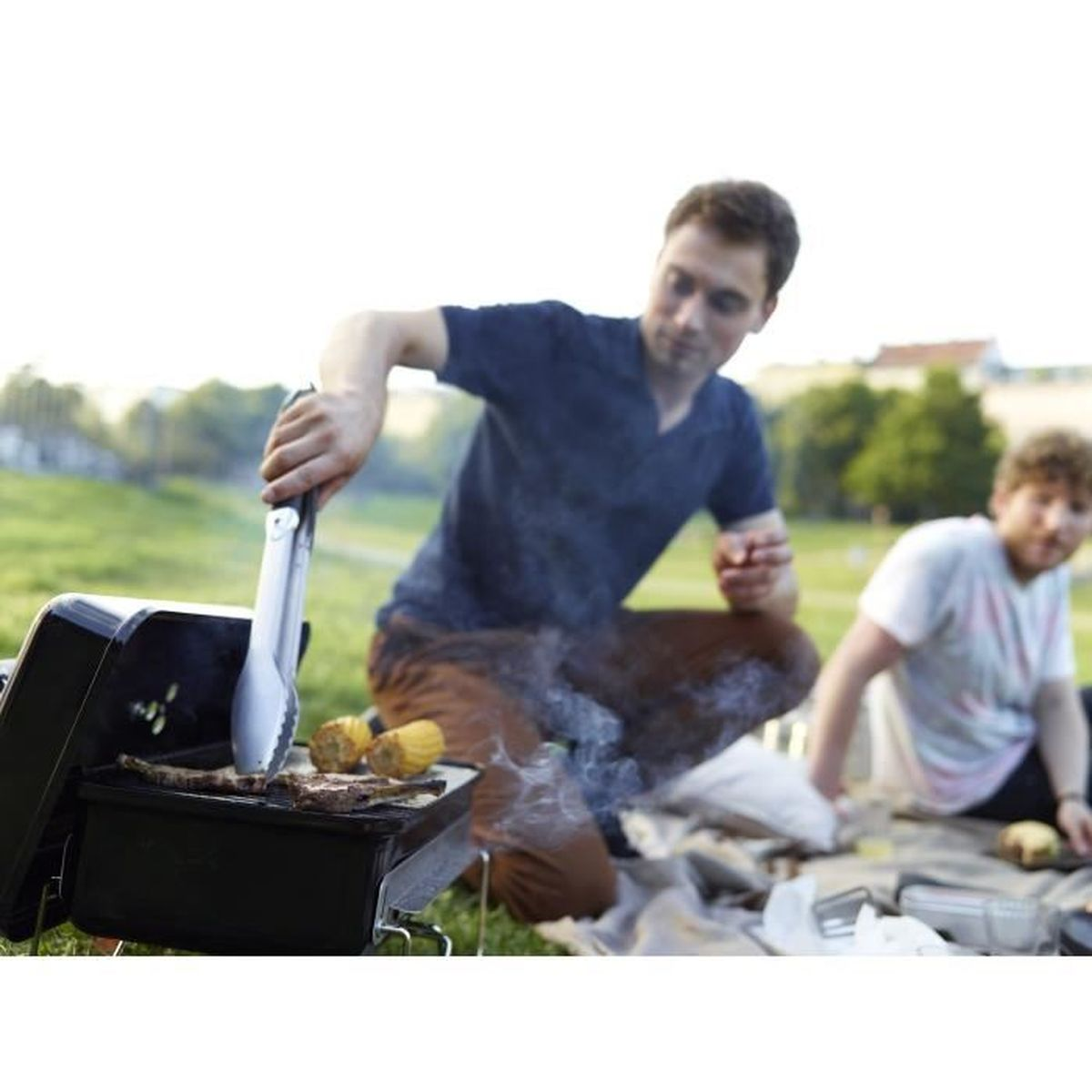 Dos hombres asando comida en una pequeña barbacoa. Un hombre usa pinzas. Fondo de hierba verde.