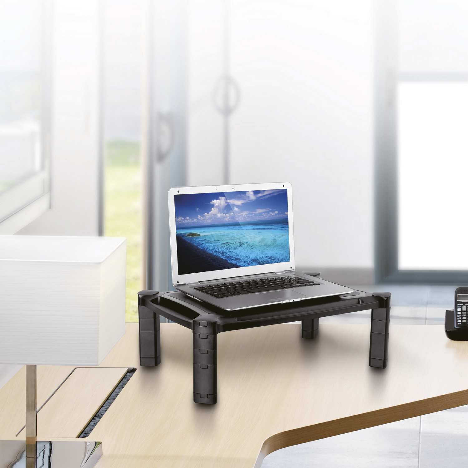 Laptop en un soporte negro, sobre un escritorio. Configuración de oficina con lámpara y ventana.