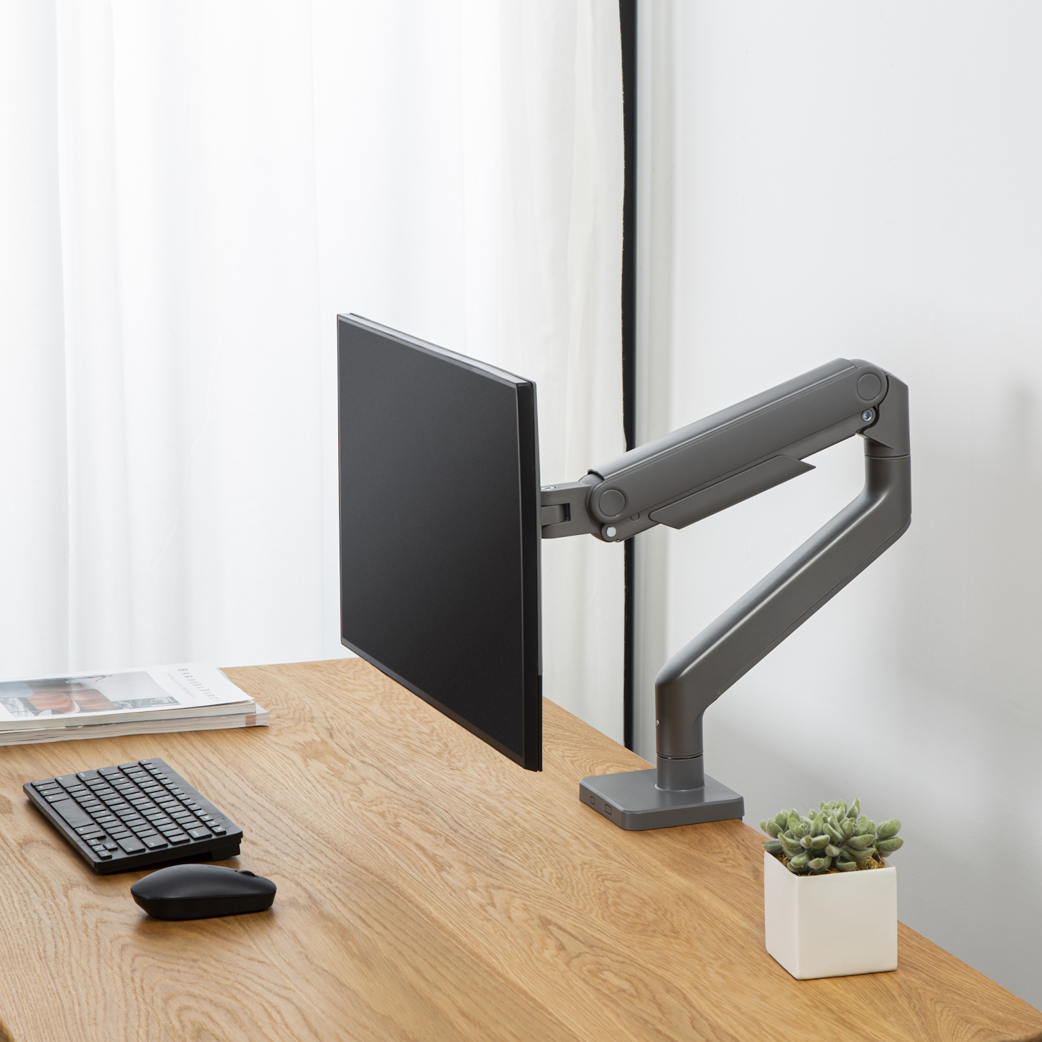 Un braccio per monitor, che tiene uno schermo nero, su una scrivania in legno con tastiera e pianta.