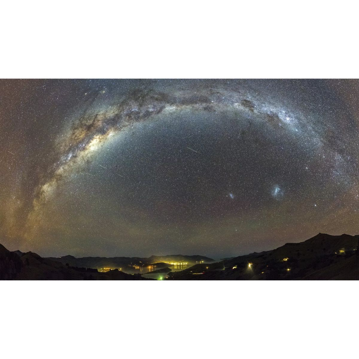 Cielo nocturno con Vía Láctea sobre montañas y luces de la ciudad. Estrellas llenan el cielo.