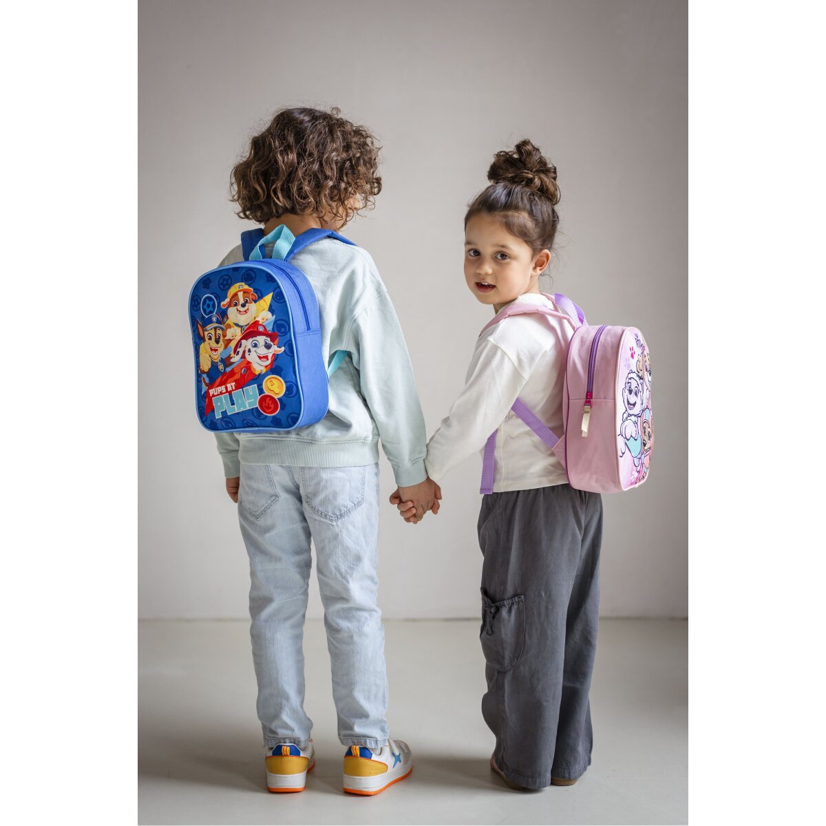 Zwei Kinder mit Paw Patrol-Rucksäcken, stehen und halten Händchen, weg von der Kamera.