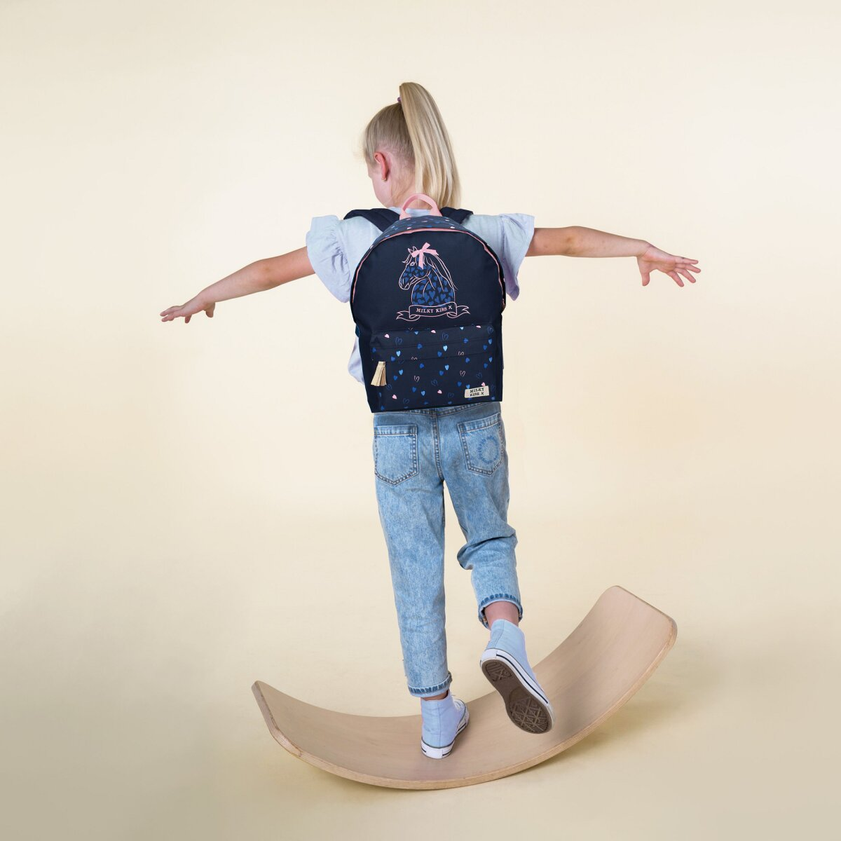 Mädchen auf einem Holz-Balance-Board, mit Rucksack, ausgestreckten Armen und hellem Hintergrund.