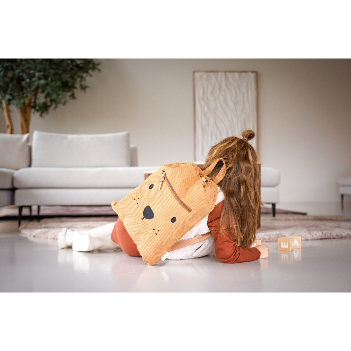 Ein Kind liegt mit einem Bären-Rucksack. Ein Holzspielzeug und ein weißes Sofa im Hintergrund.