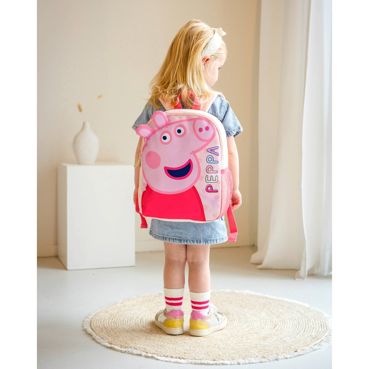 Blondes Mädchen mit Peppa Pig-Rucksack, das sich abwendet. Sie steht auf einem runden Teppich.