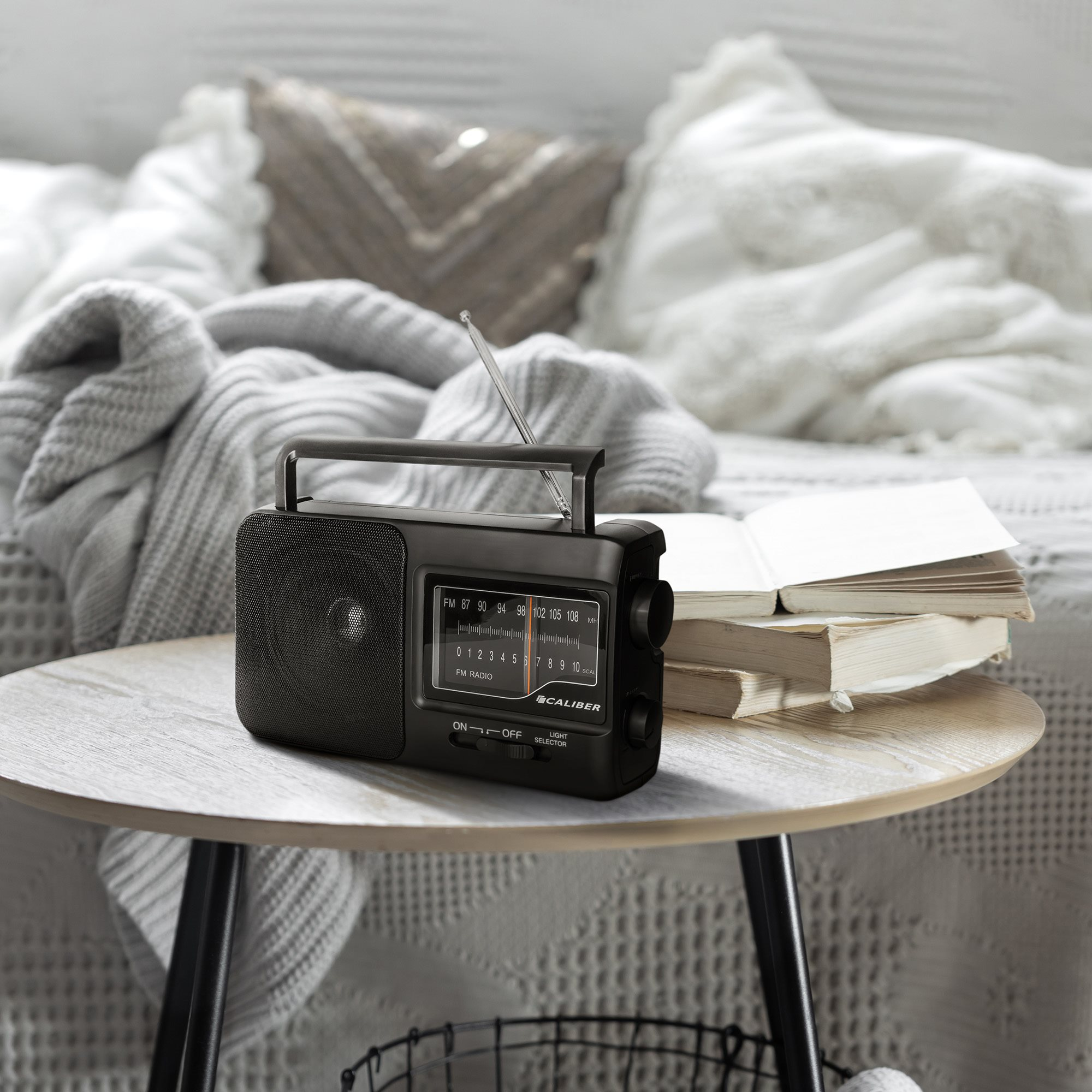 A black radio sits on a wooden table with a stack of books. A bed and pillows are in the background.