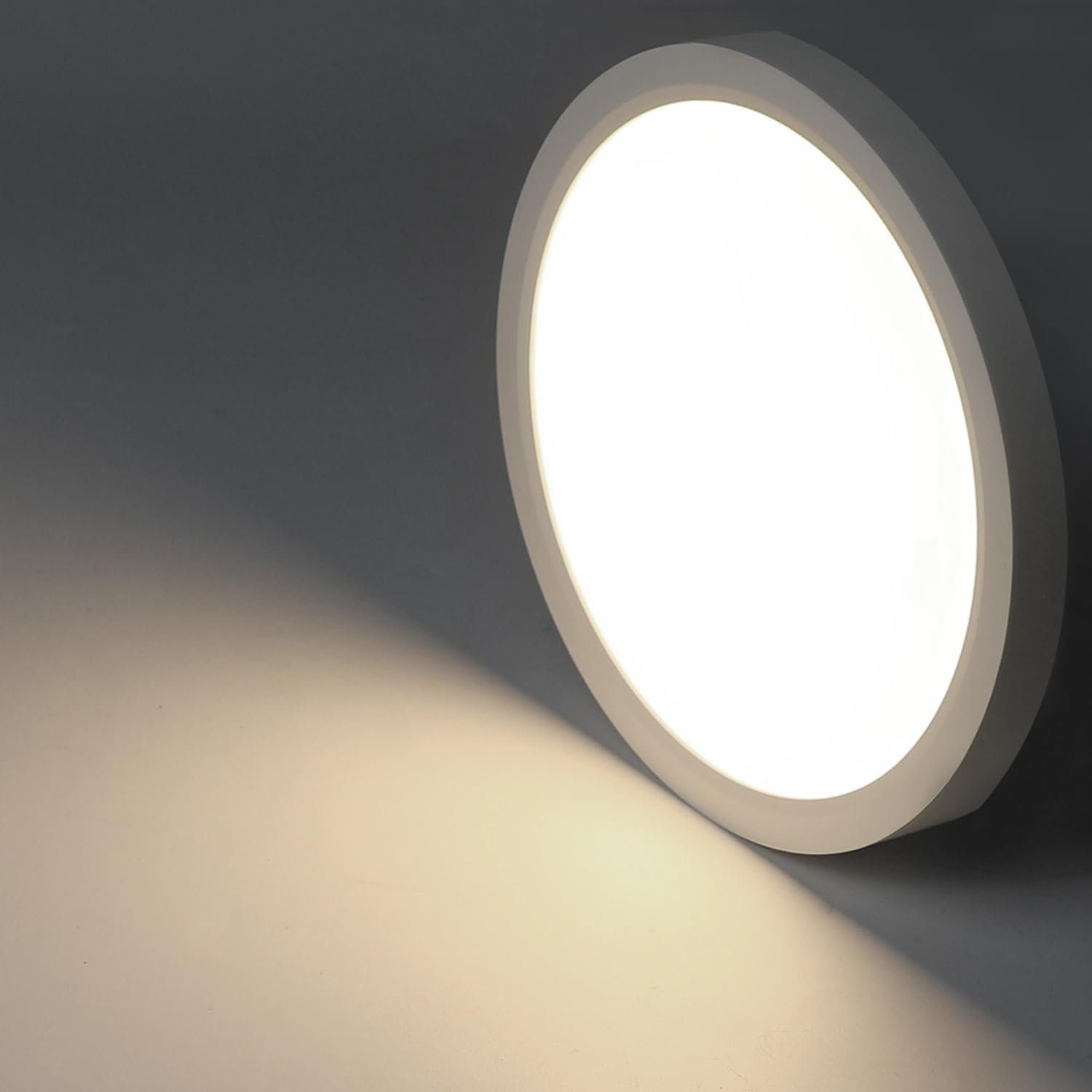 Una plafoniera bianca, rotonda e illuminata contro un muro grigio scuro, che proietta un bagliore in basso.