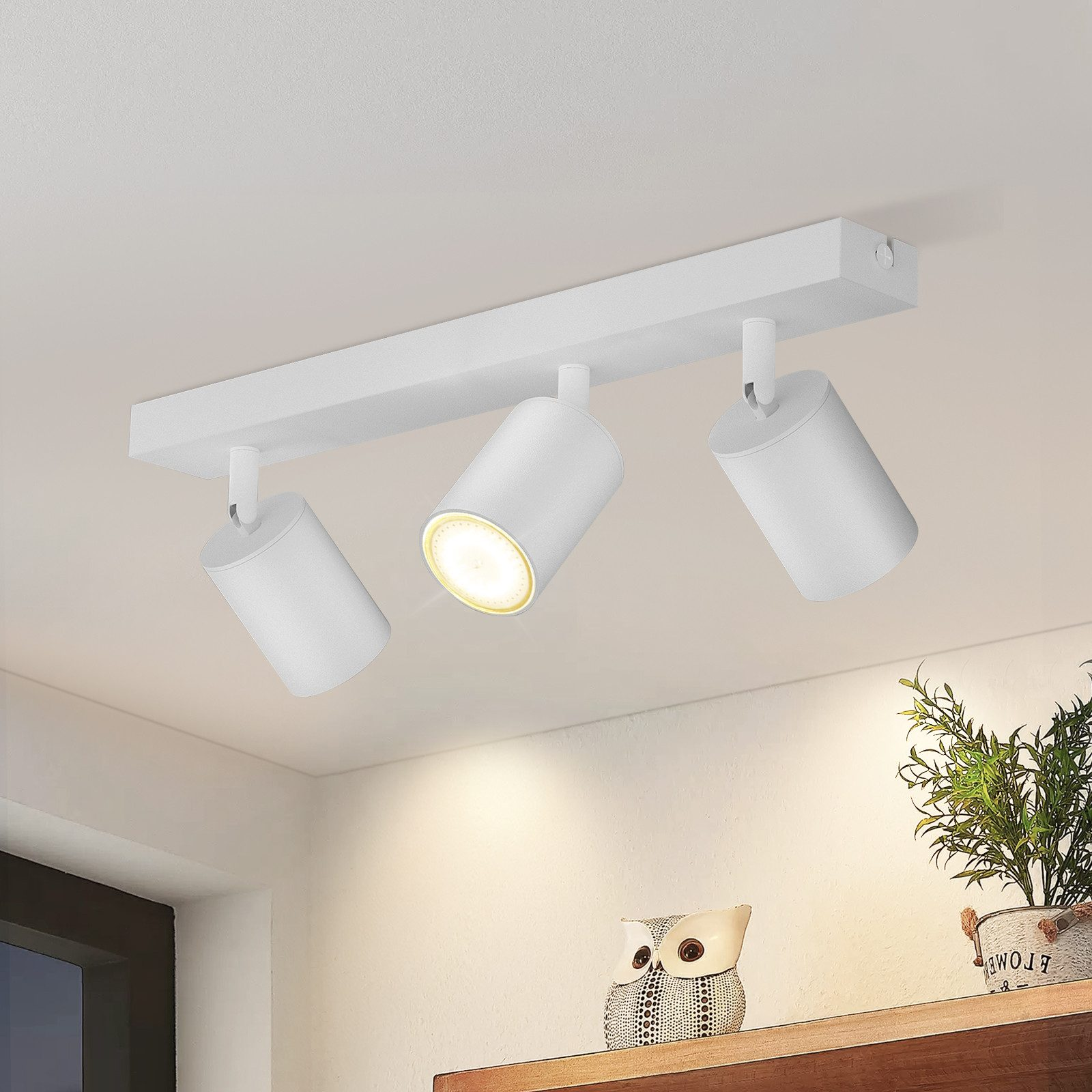 Tre faretti bianchi su una barra bianca montata a soffitto. Lo sfondo include una mensola con oggetti.