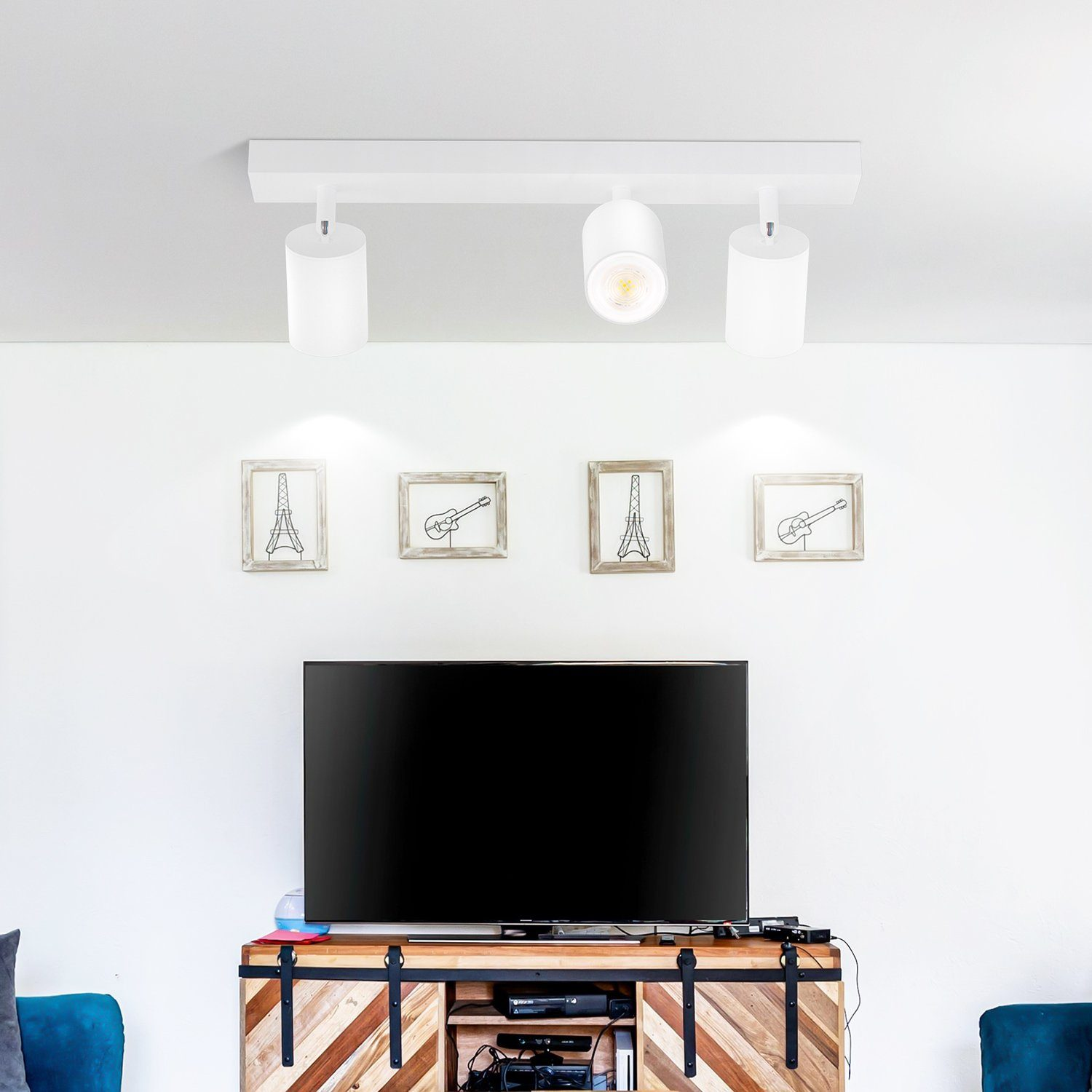 Tre faretti bianchi sul soffitto. Sotto, una TV e opere d'arte. Muri bianchi e mobile in legno.