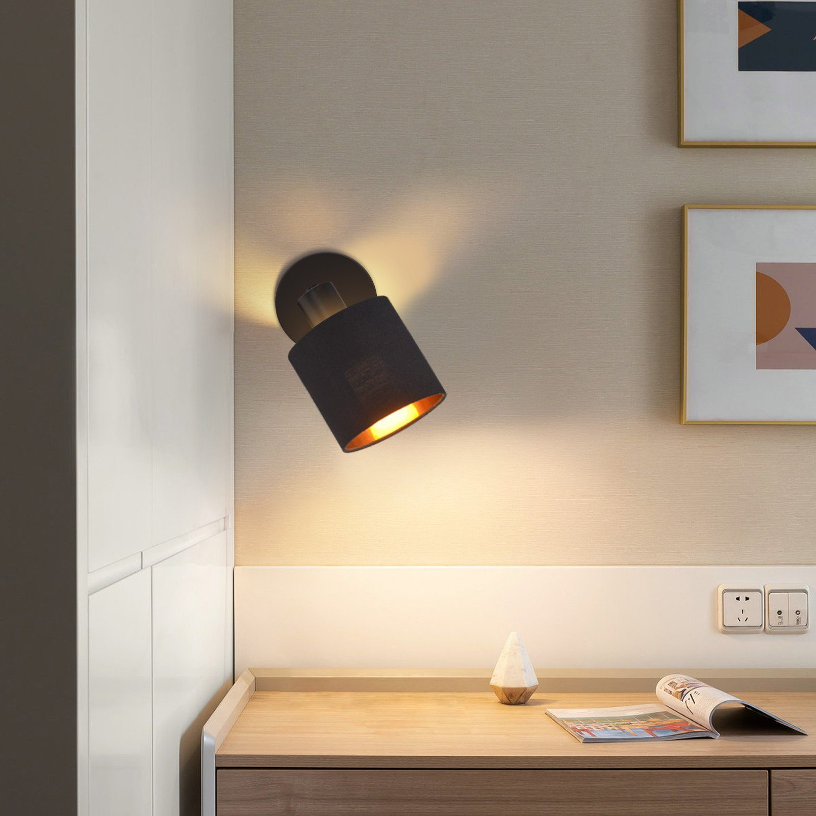 Lampada da parete nera con interno dorato montata su parete beige. Due quadri sul muro.