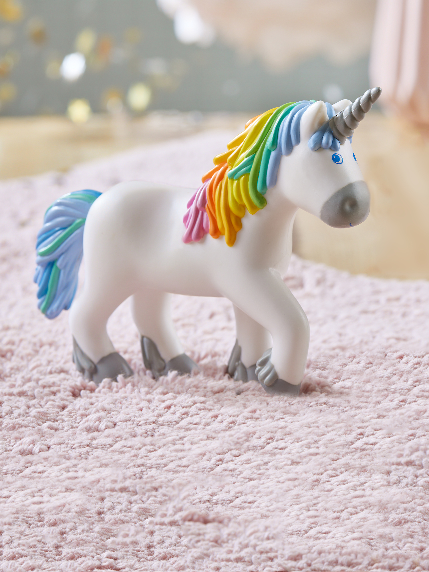 Weißes Einhornspielzeug mit Regenbogenmähne und -schwanz auf einem rosa Teppich. Es hat ein silbernes Horn und graue Hufe.