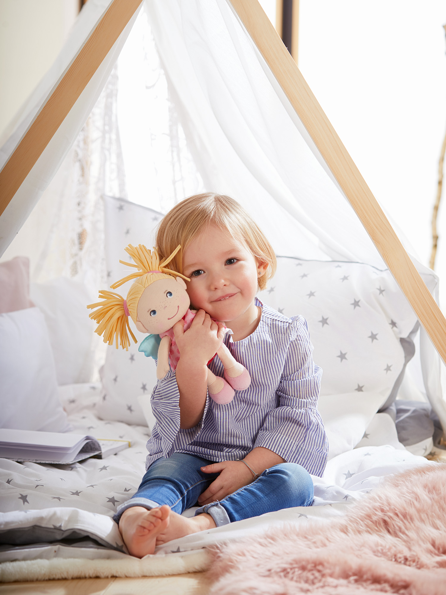 Kleines Mädchen, das eine Puppe hält und auf einem Bett sitzt. Der Hintergrund ist weiß.
