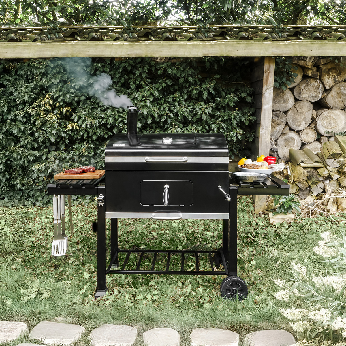 Zwarte buitengrill op gras. Inclusief een bijzettafel met eten, gereedschap en een schoorsteen. Houtopslag op de achtergrond.
