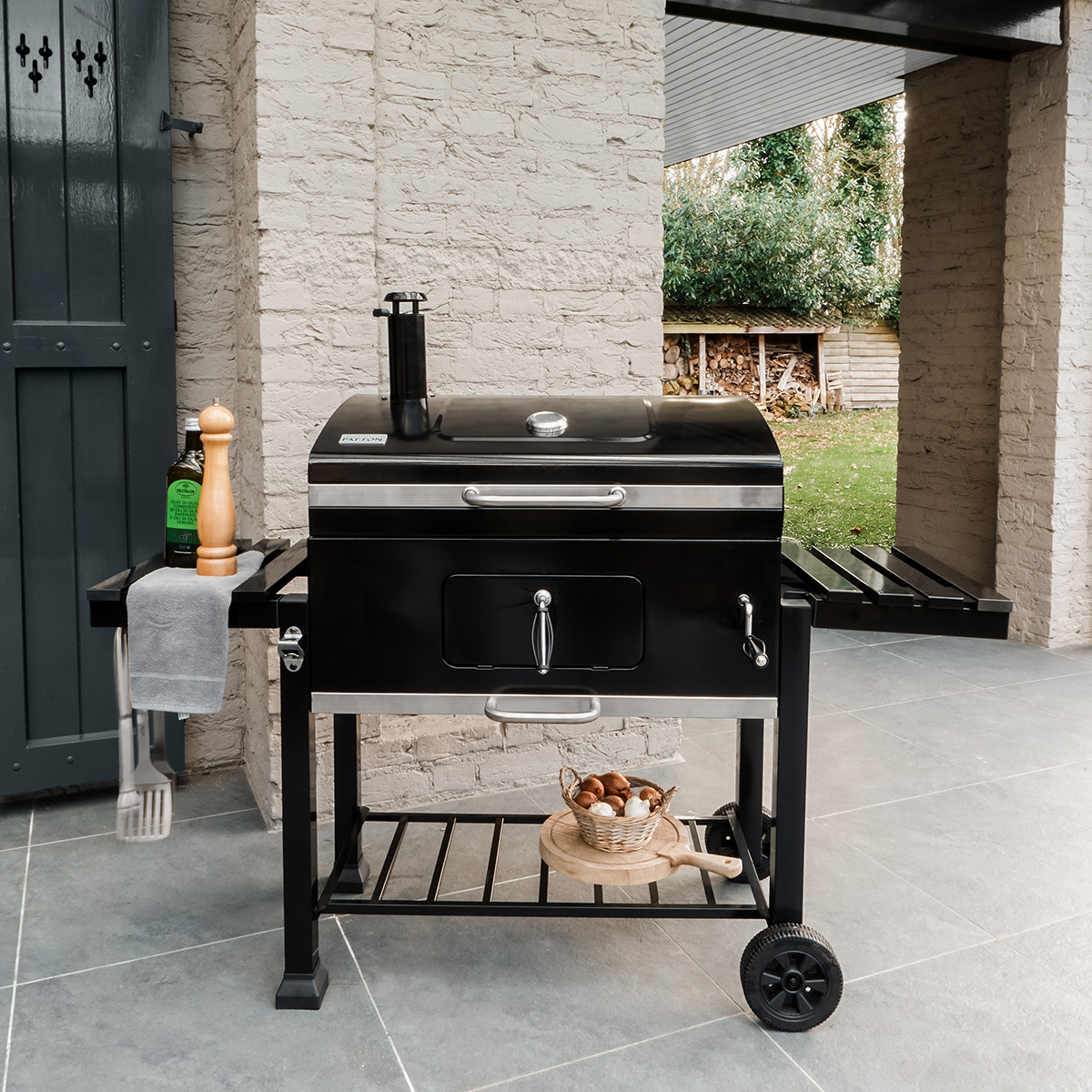 Zwarte buitengrill met zijplank. Bakstenen muur en buitenomgeving. Hout en keukengerei zichtbaar.