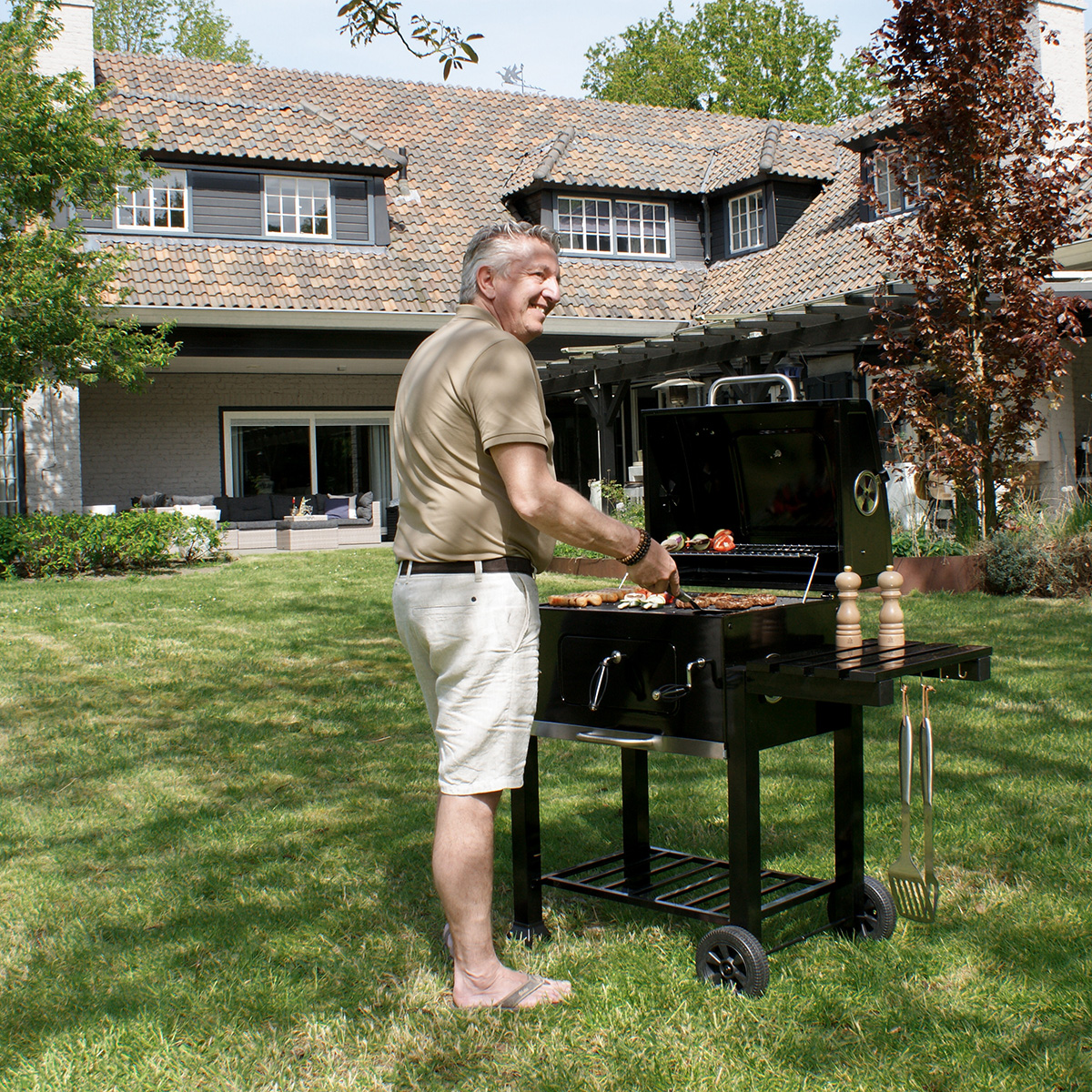 Ein Mann grillt Essen auf einem schwarzen Grill im Hinterhof, trägt ein beiges Hemd und weiße Shorts. Sonnig.