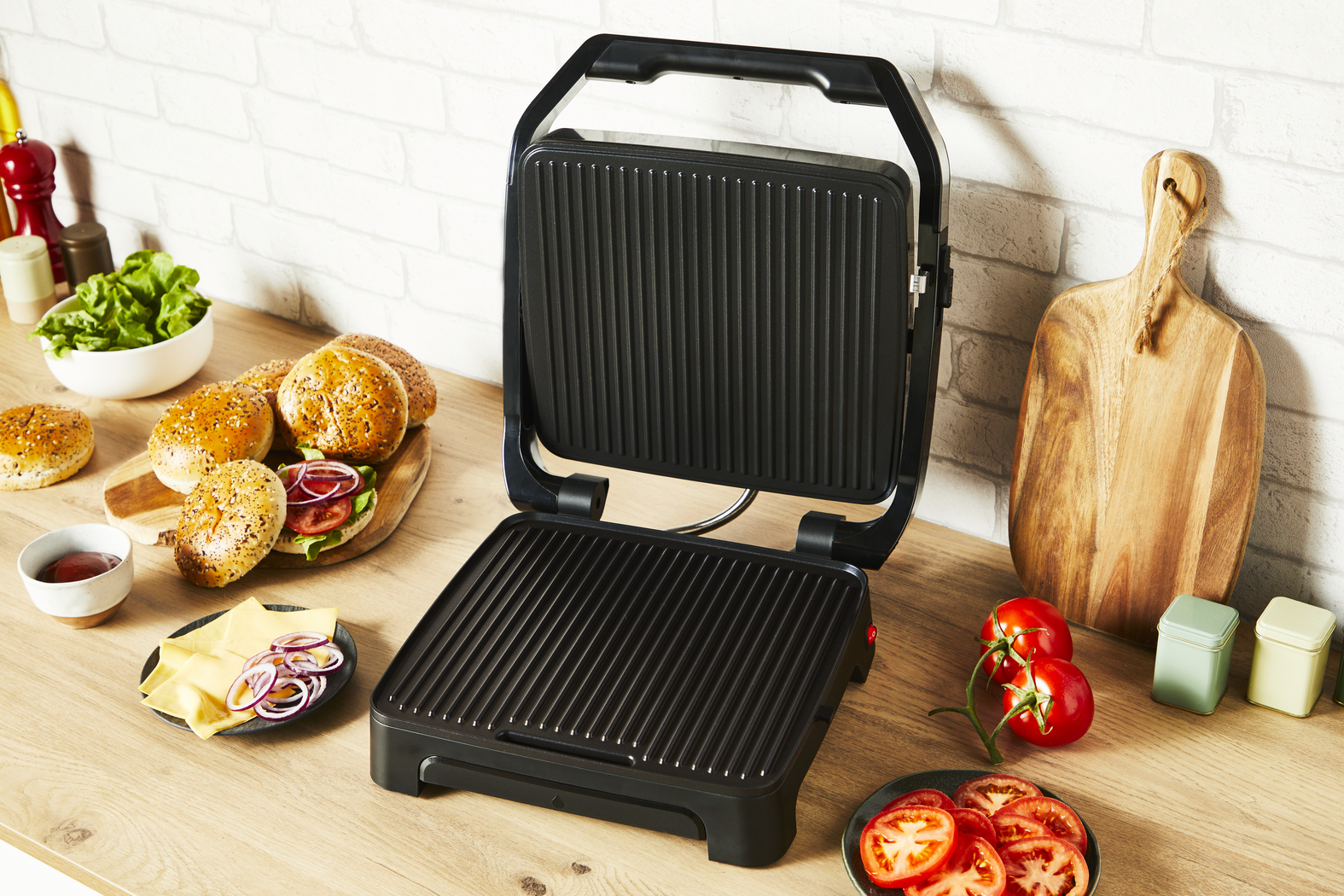 Ein offener schwarzer Grill mit Burgern und Sandwichzutaten auf einer Holz-Arbeitsplatte.