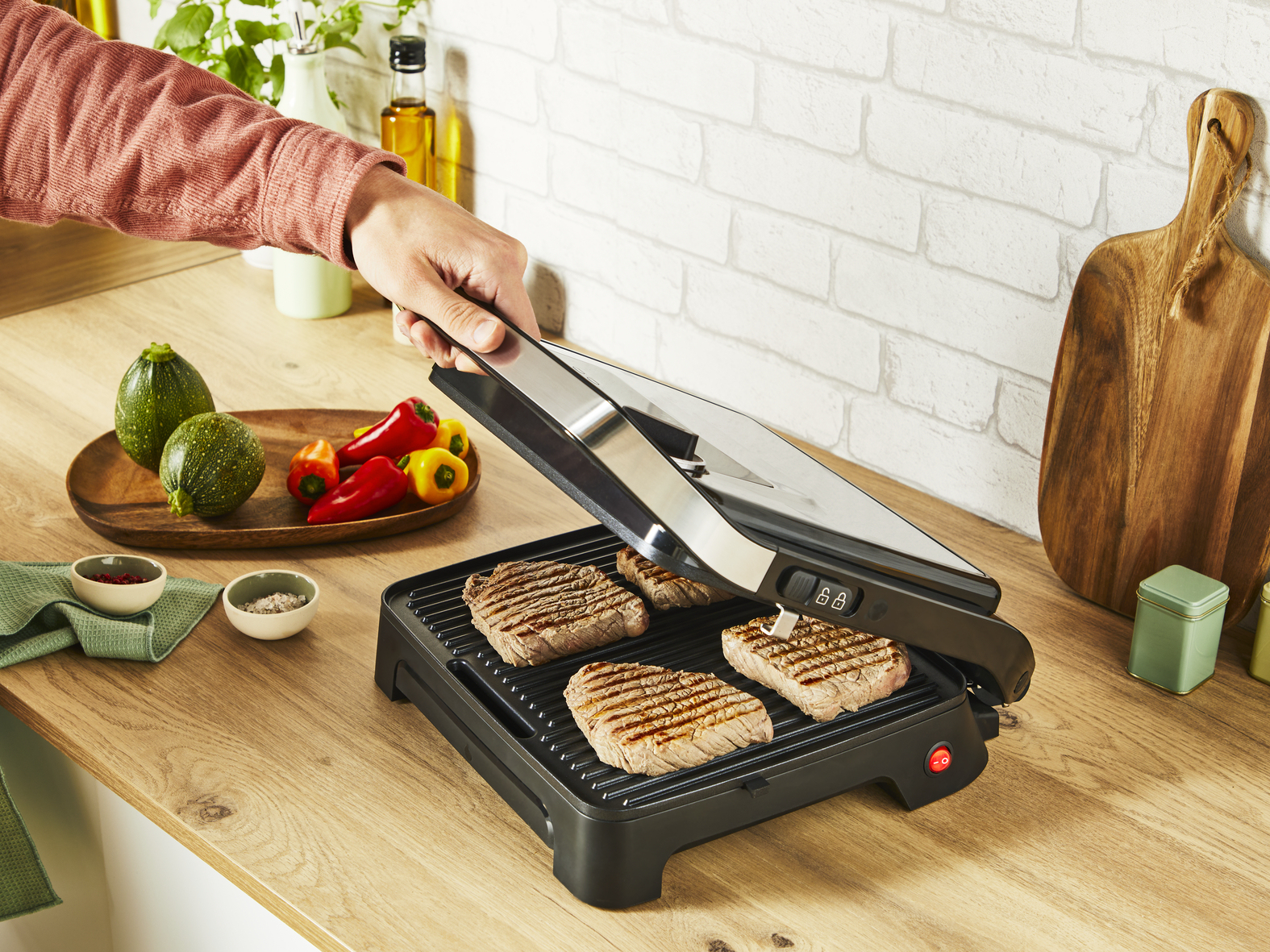 Eine Person grillt Steaks auf einem schwarzen Grill, Gemüse auf einer Holz-Arbeitsplatte.