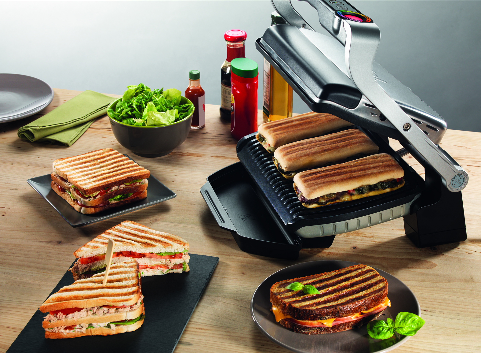 Eine Panini-Presse mit mehreren Sandwiches, Salaten und Gewürzen auf einem Holztisch.