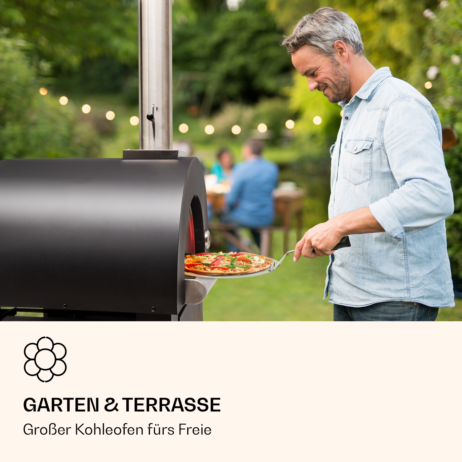 Ein Mann legt eine Pizza in einen schwarzen Outdoor-Ofen. Im Hintergrund sind weitere Personen und Lichter.