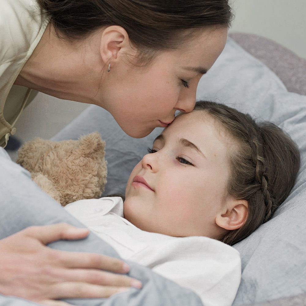Matka całuje dziecko w czoło, leżące w łóżku z misiem.