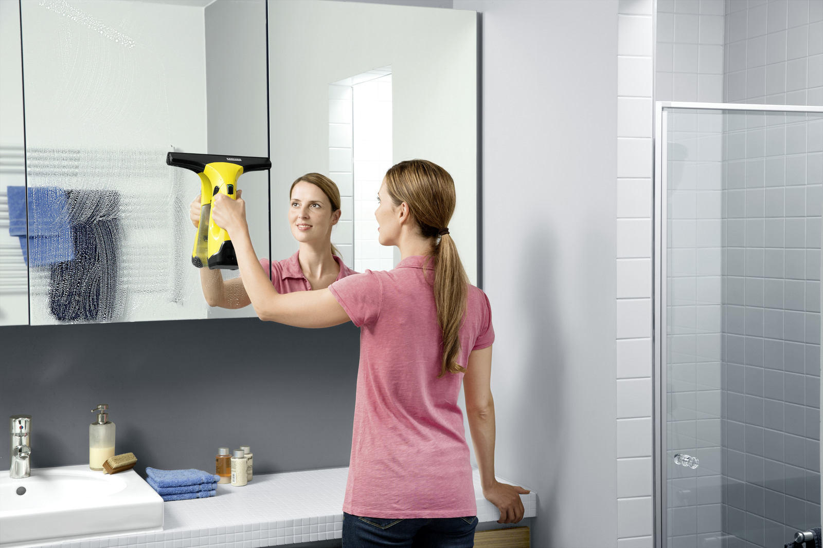 Una donna pulisce uno specchio in un bagno con un tergivetro giallo, con il suo riflesso.