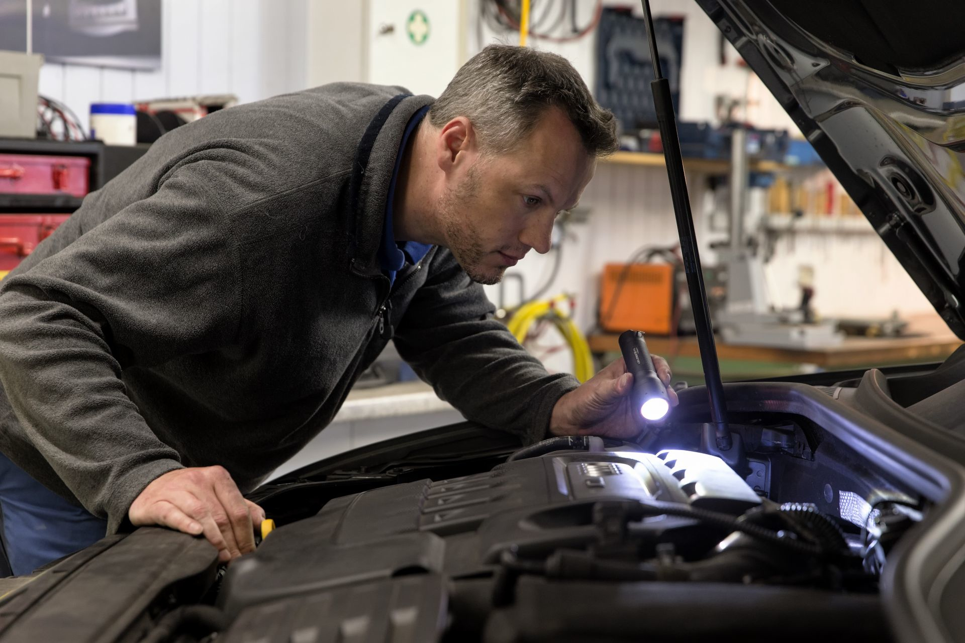 Mężczyzna w garażu, ogląda silnik samochodu z latarką.