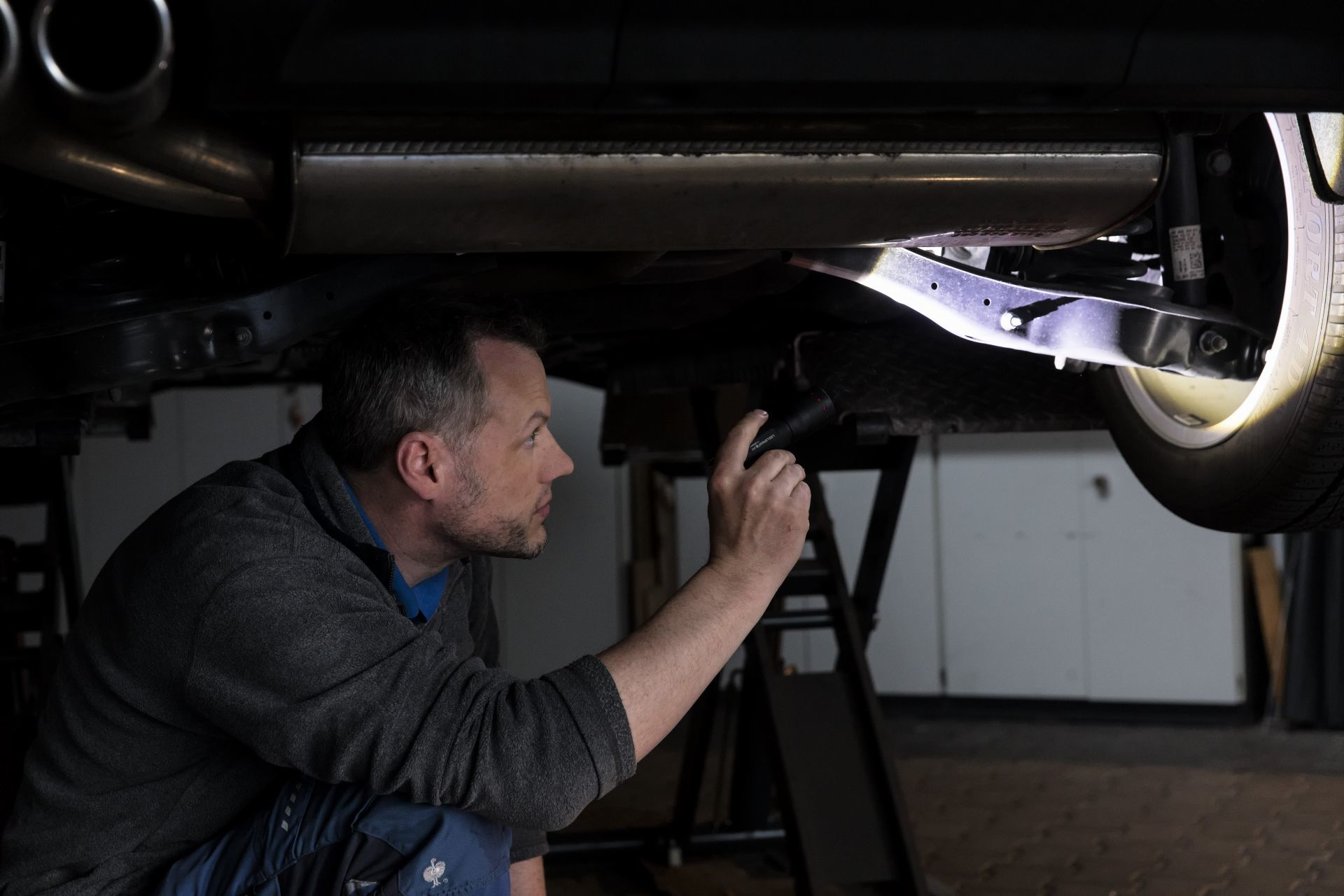 Mężczyzna w szarych ubraniach sprawdza spód samochodu latarką w otoczeniu garażu.