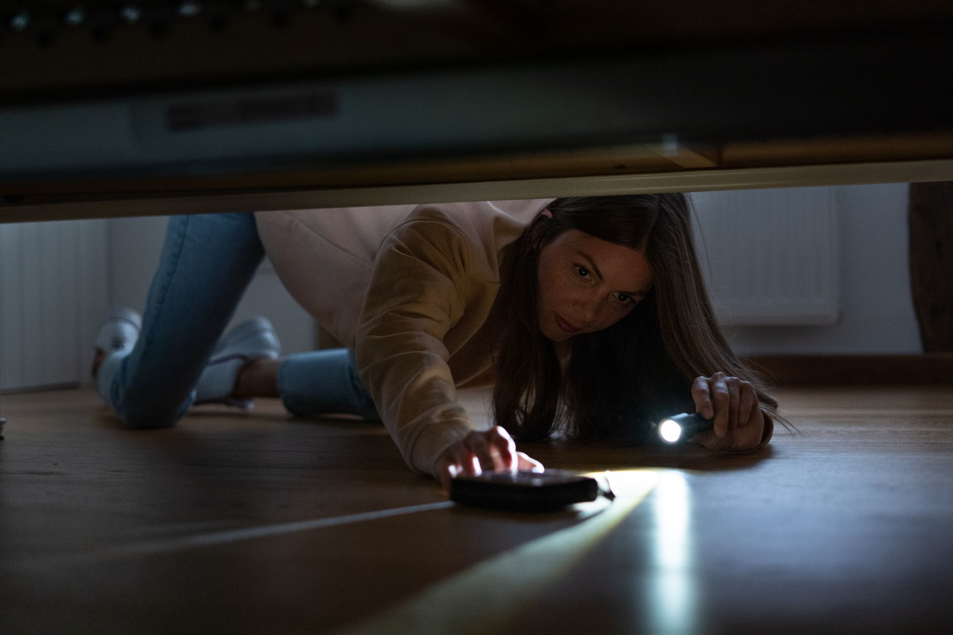 Kobieta z latarką szuka telefonu pod łóżkiem na drewnianej podłodze.