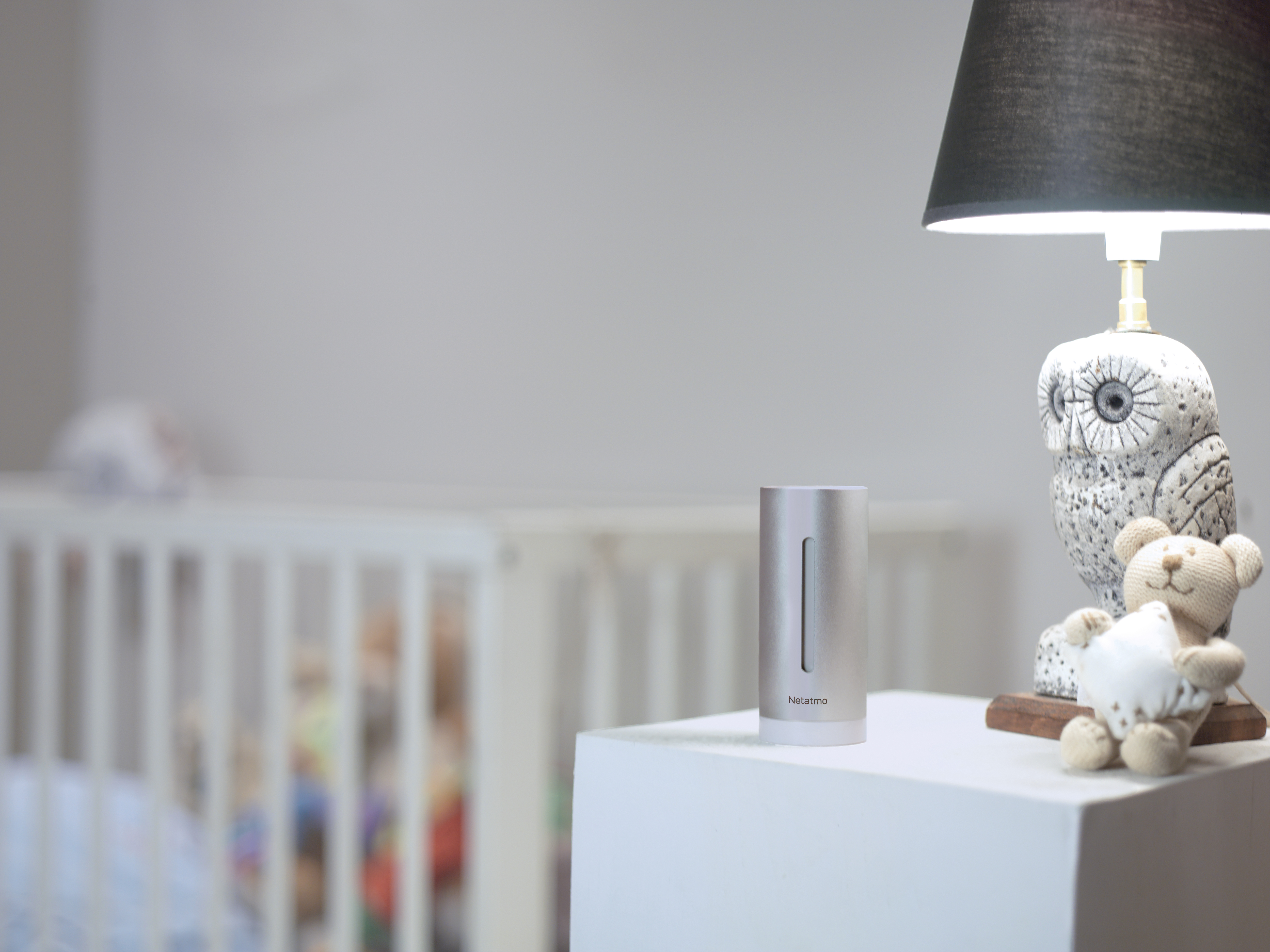 Urządzenie Netatmo na białym stole, obok lampy i misia. W tle kołyska.