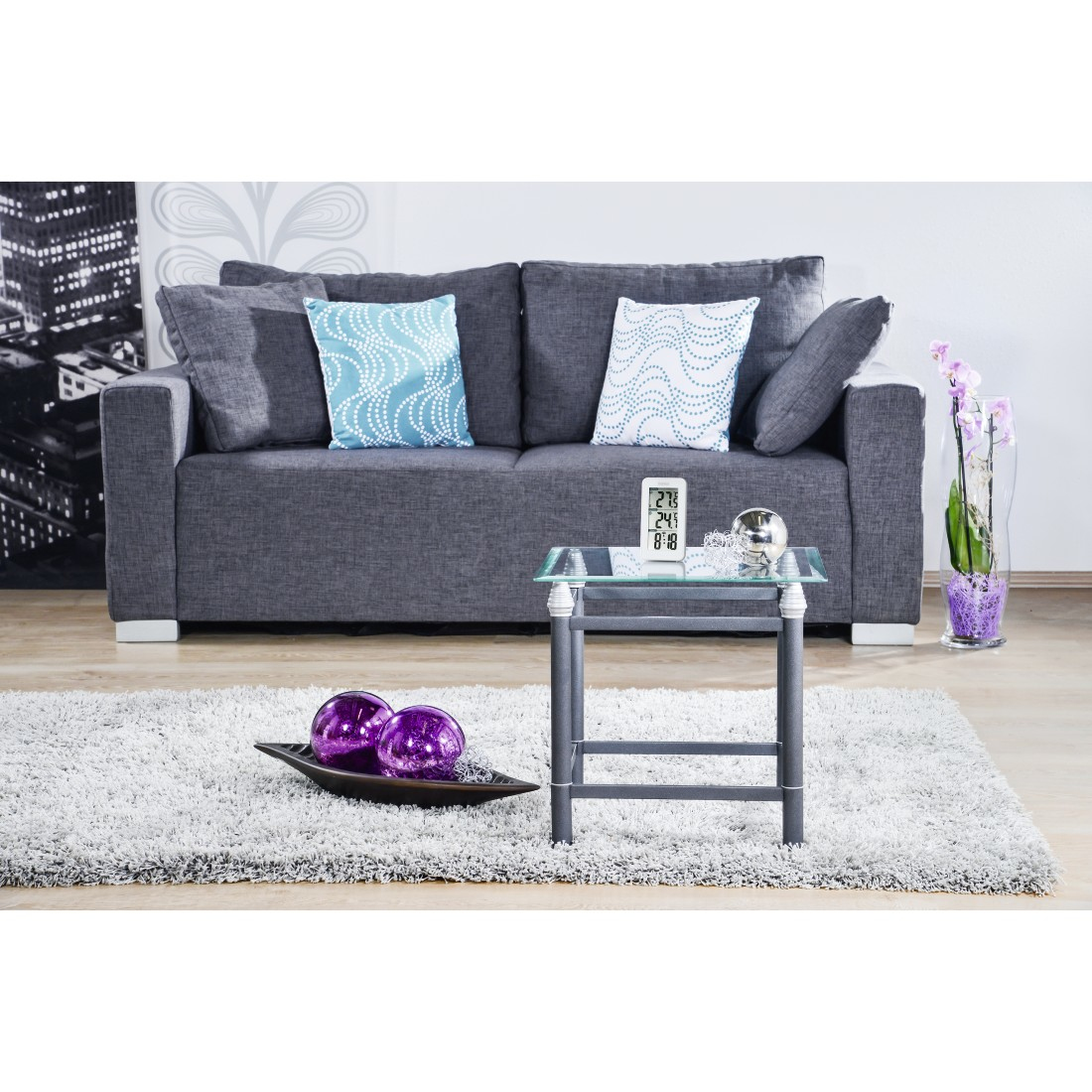 Szara sofa z niebieskimi poduszkami, szklany stolik, fioletowe kule i puszysty dywan.