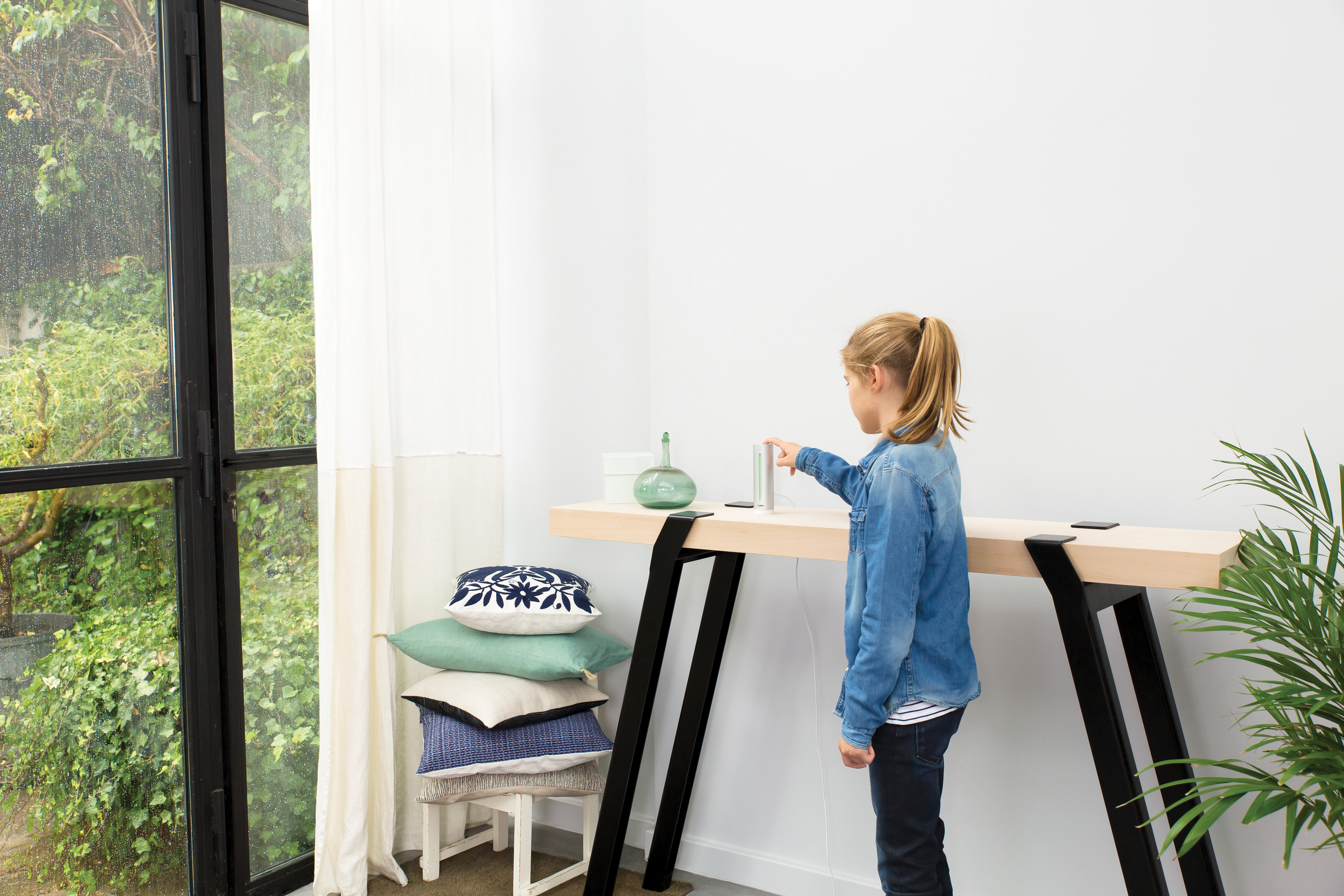 Dziewczynka dotyka srebrnego przedmiotu na drewnianym stole. Poduszki i rośliny.