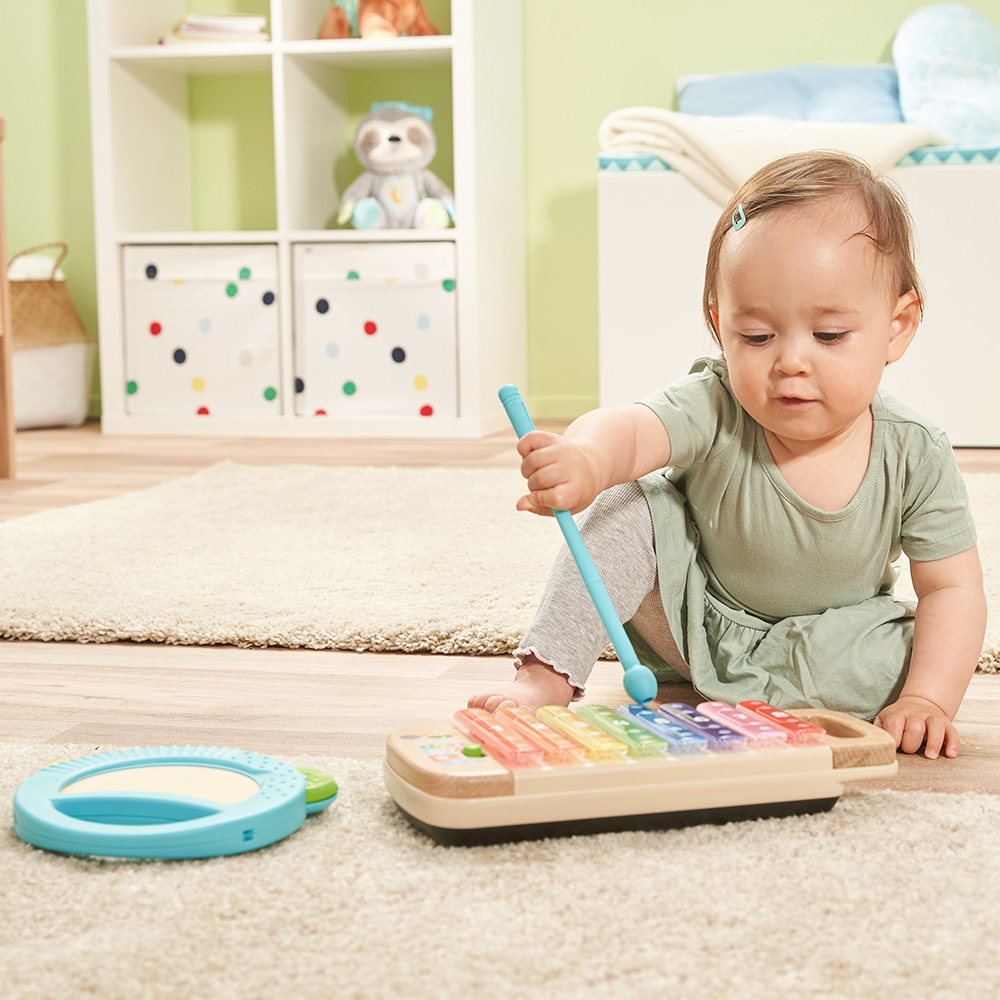 Ein Baby spielt mit einem Schlägel Xylophon, in der Nähe einer Trommel, auf einem Teppichboden.
