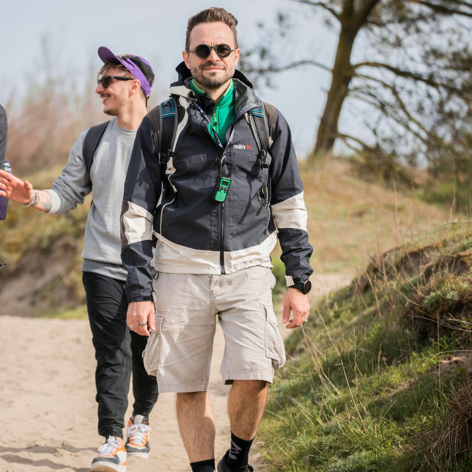 Trzech mężczyzn idących piaszczystą ścieżką. Jeden w centrum, w okularach i kurtce.