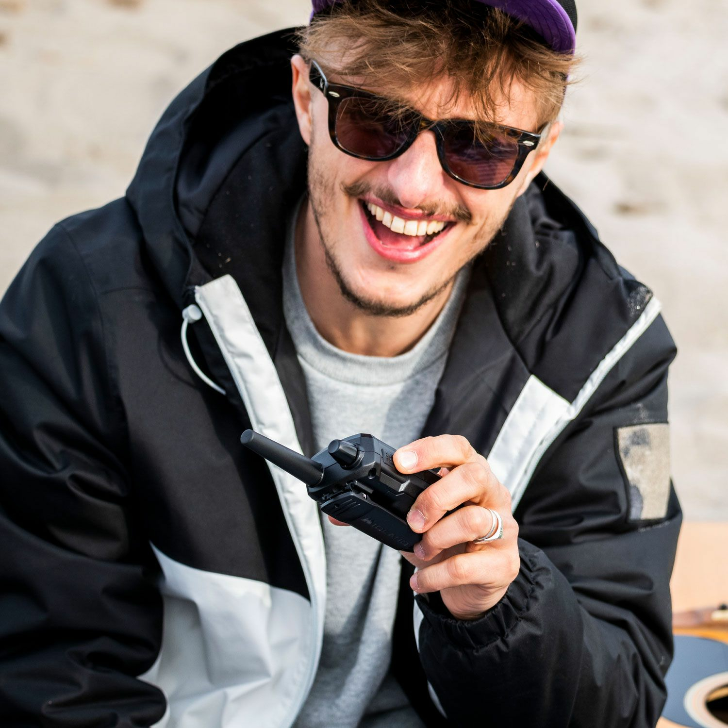 Uśmiechnięty mężczyzna w okularach przeciwsłonecznych trzyma krótkofalówkę. Ma na sobie czarną kurtkę i fioletową czapkę.
