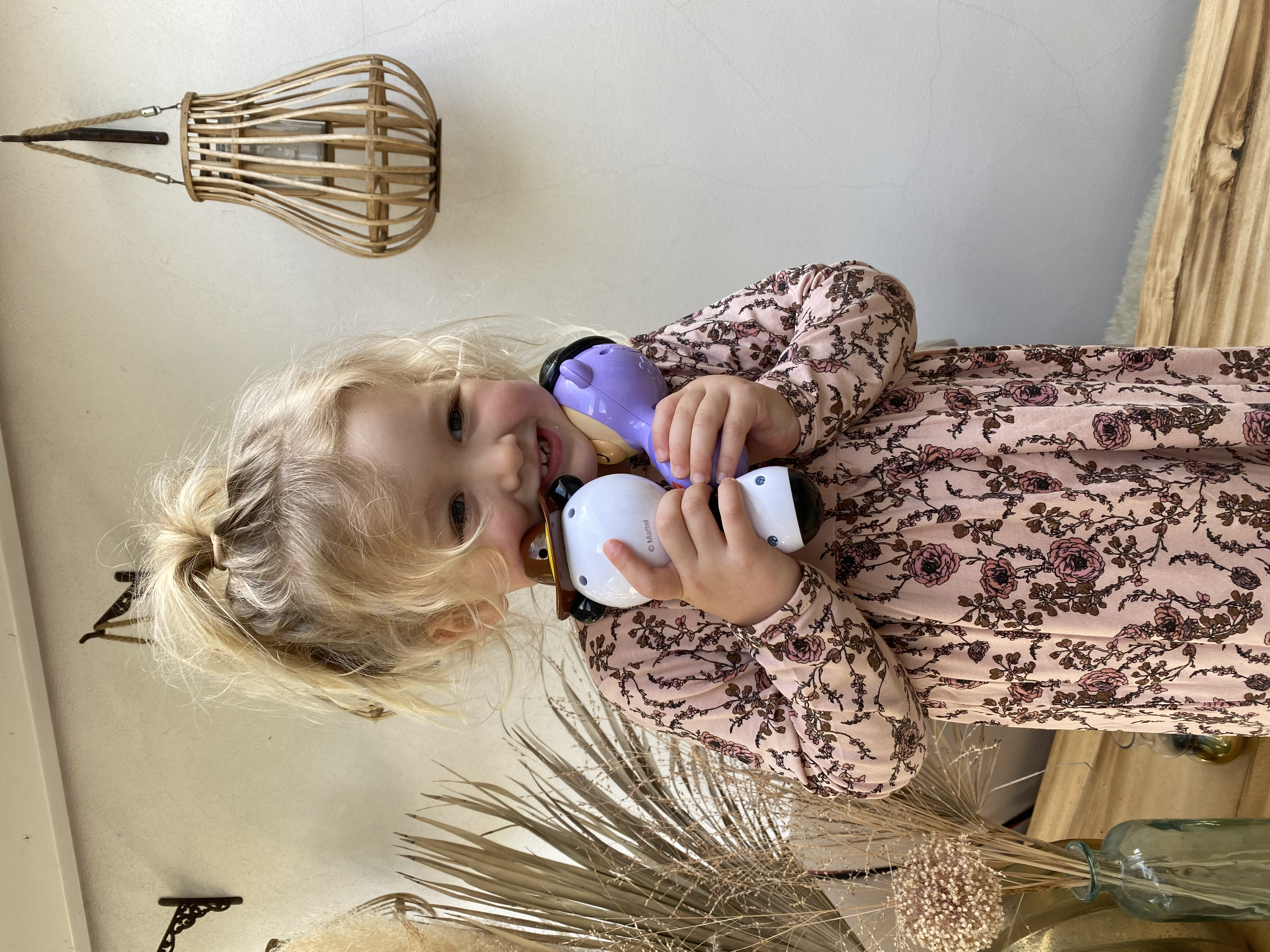 Niña pequeña sosteniendo dos juguetes, sonriendo, con vestido floral, fondo casual.