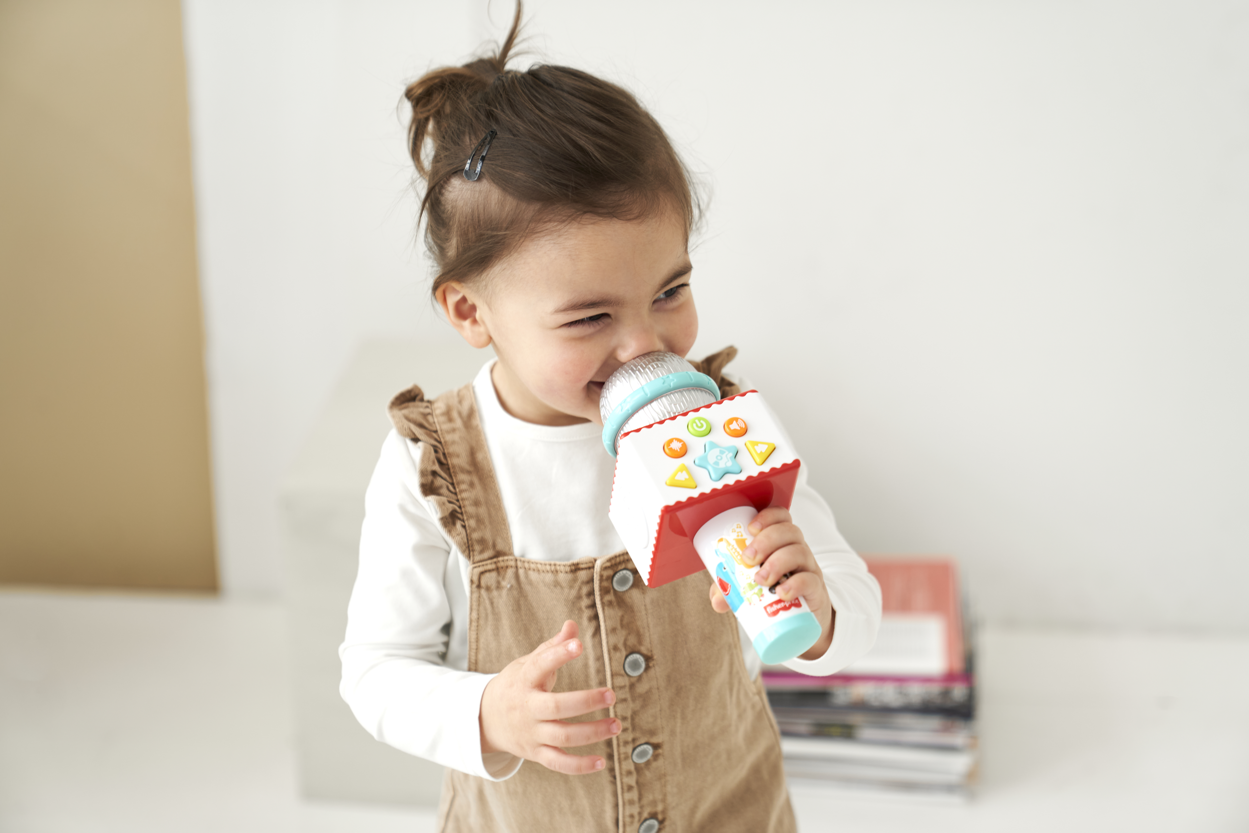 Mała dziewczynka trzyma zabawkę mikrofon, uśmiechając się. Ubrana w brązowy kombinezon.