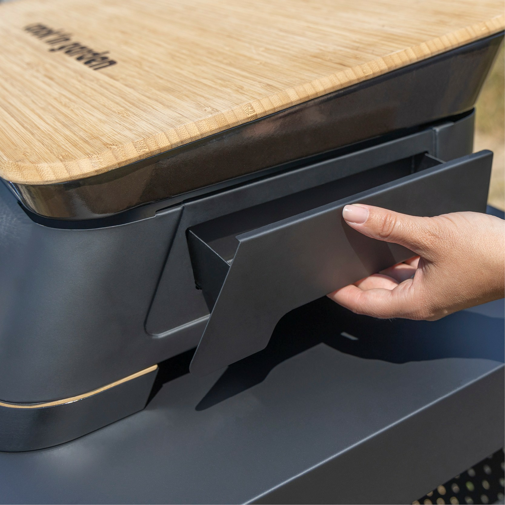 Ręka otwiera szufladę grilla zewnętrznego, z drewnianym blatem i czarną ramą.