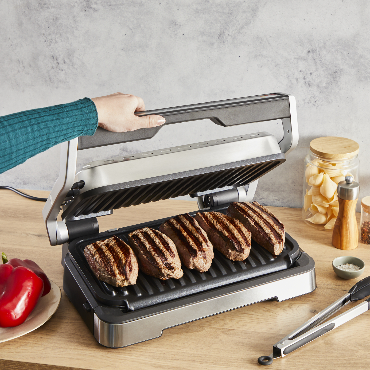 Grill mit sechs Steaks, die gegart werden. Eine Hand hält den Deckel. Zangen und Paprika sind in der Nähe.