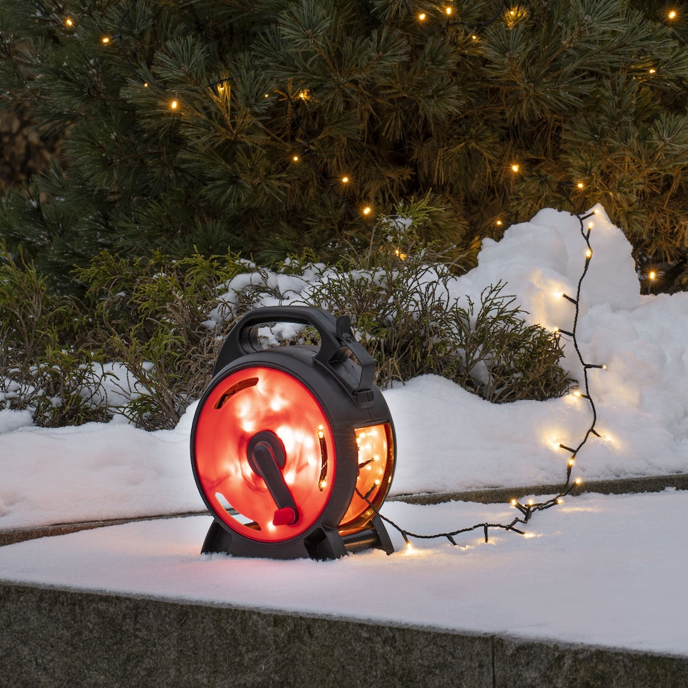 Eine schwarze Kabeltrommel mit Lichtern im Schnee neben einem Weihnachtsbaum.