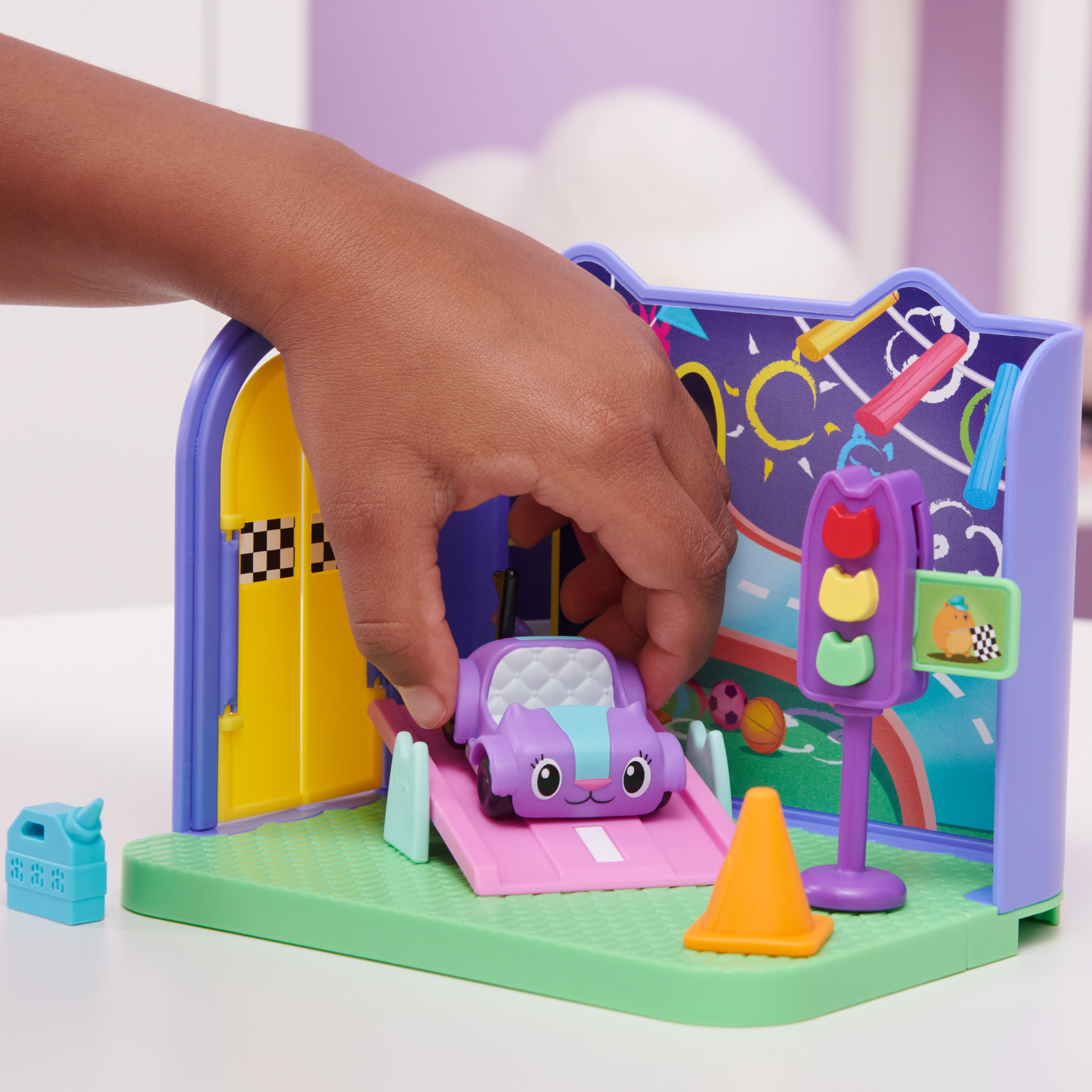 Kinderhand, die ein Spielzeugauto auf eine lila Rampe stellt. Die Szene beinhaltet eine Ampel und eine kleine Garage.