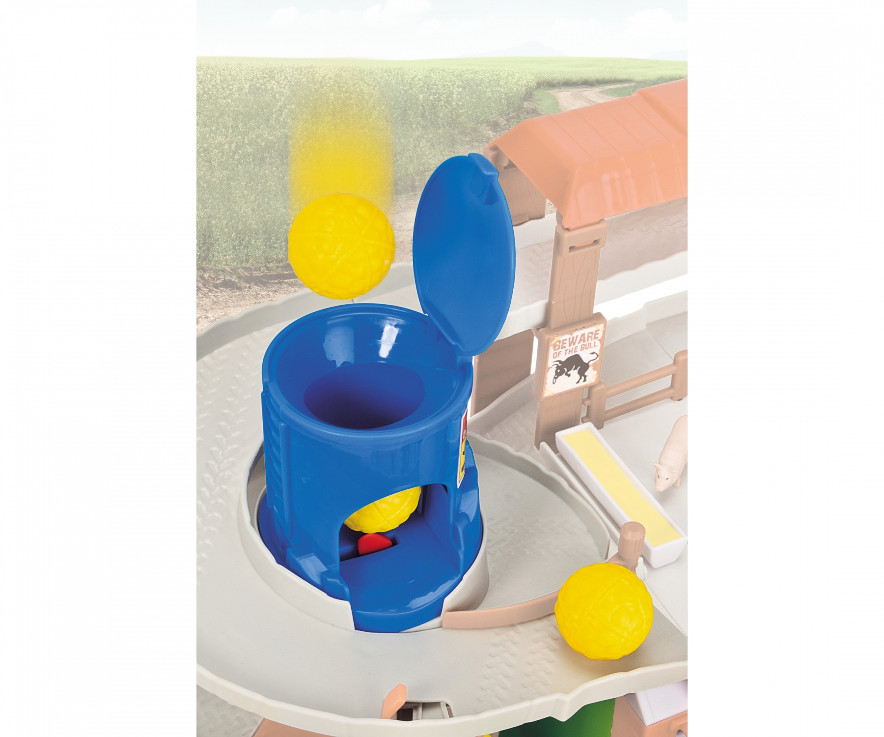 Eine Spielzeug-Bauernhofszene mit einem blauen Silo, das gelbe Bälle ausgibt. Ein Schwein und ein Bauernhofgebäude sind ebenfalls sichtbar.