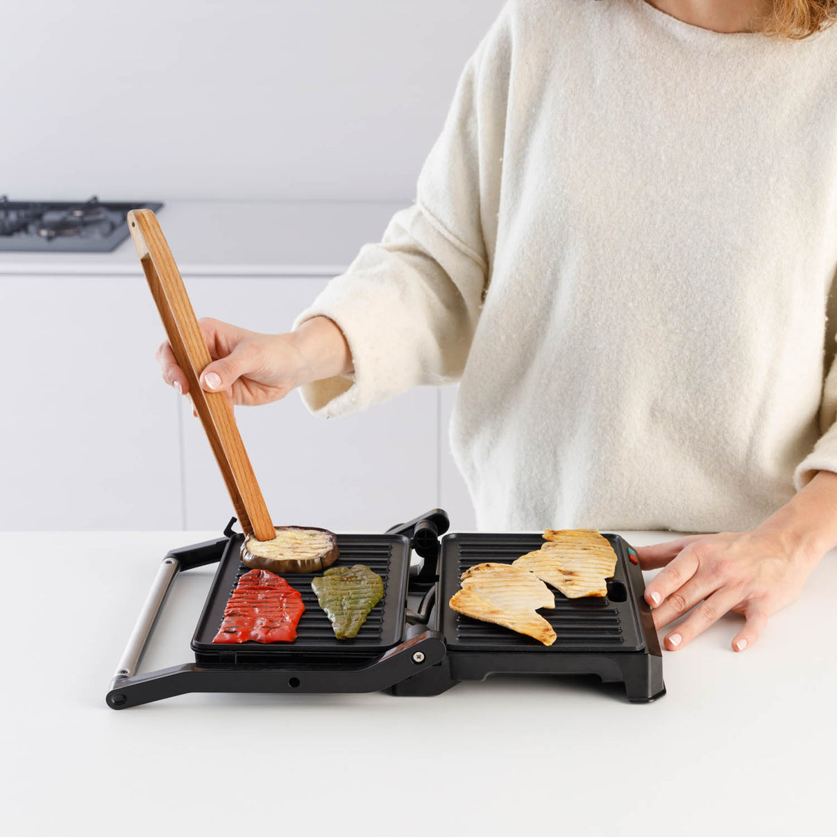 Osoba używa drewnianych szczypiec do przewracania jedzenia na czarnym grillu. Warzywa są grillowane.