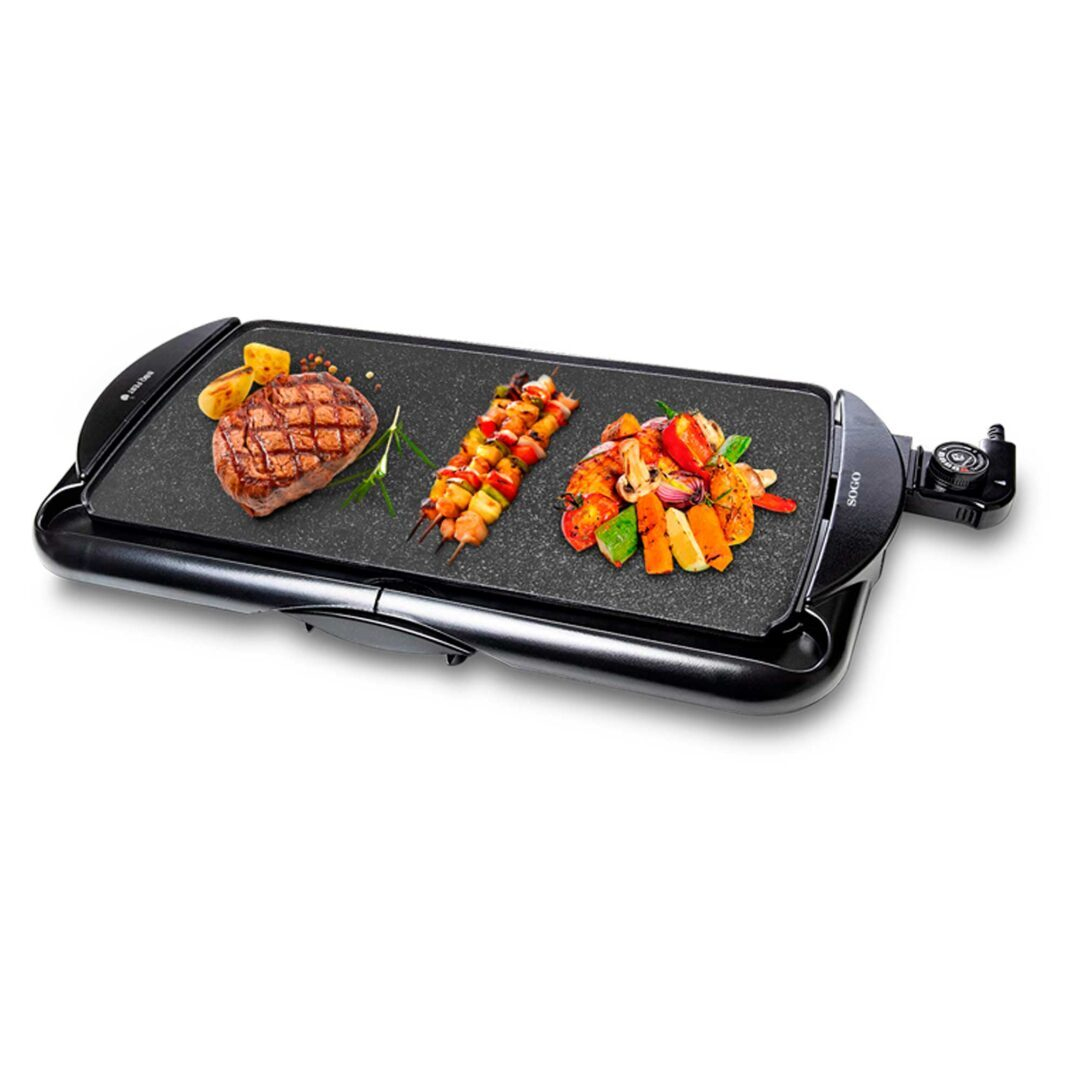 Una parrilla eléctrica negra con bistec cocido, brochetas y verduras. La parrilla está sobre una superficie blanca.