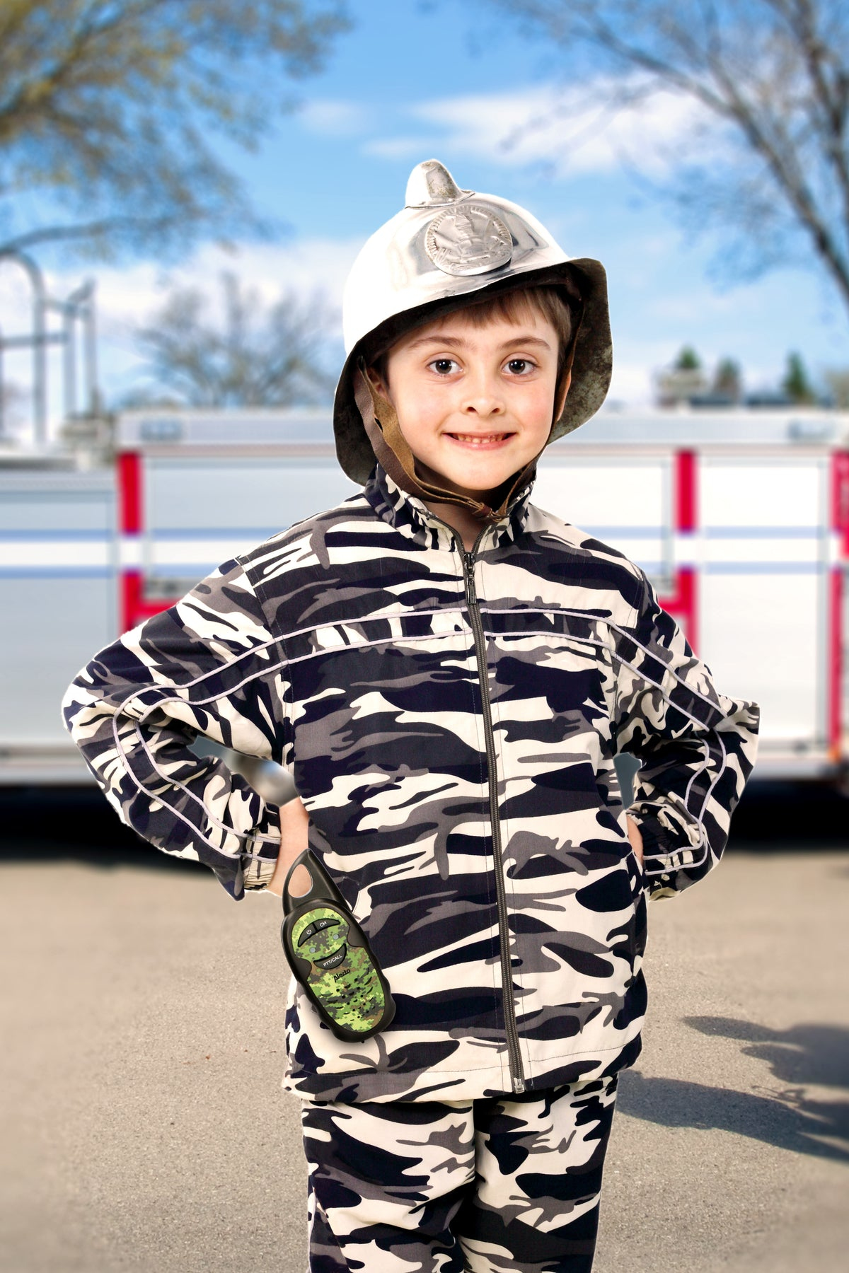 Chłopiec w stroju strażaka uśmiecha się. W tle rozmyty wóz strażacki i drzewa.