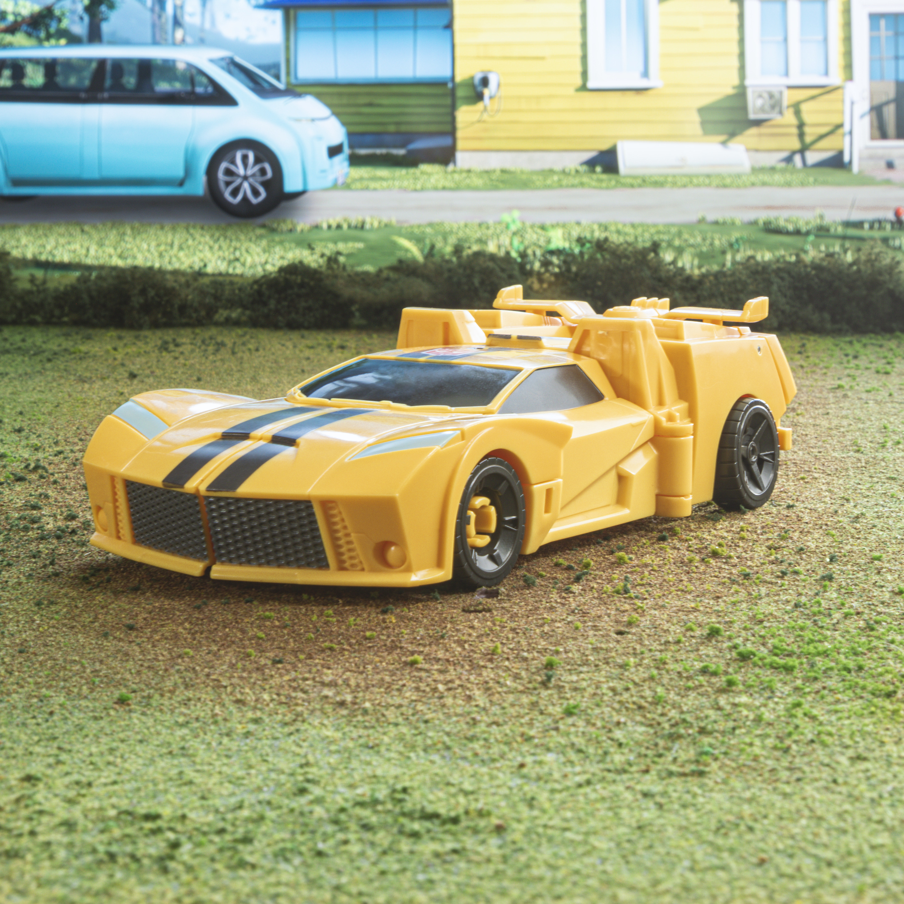 Coche de juguete amarillo sobre hierba, con coche azul detrás.