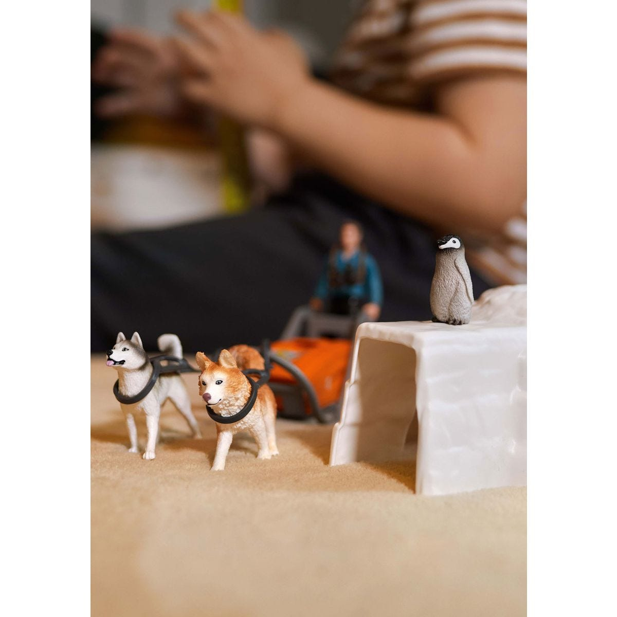 Des chiens husky jouets tirent un traîneau avec une personne, un pingouin et un igloo sur une surface beige.