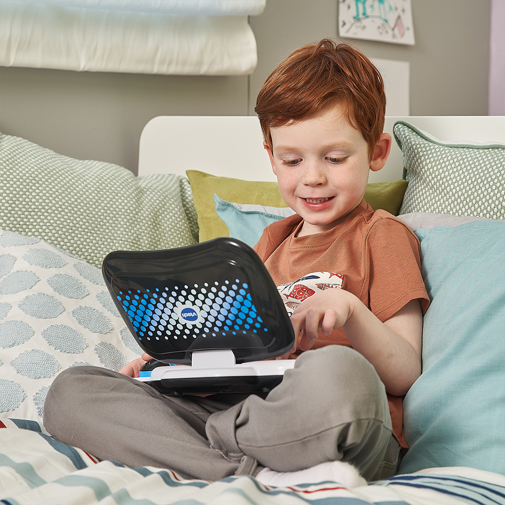 Ein kleiner Junge sitzt auf einem Bett und spielt mit einem Spielzeug-Laptop und lächelt.