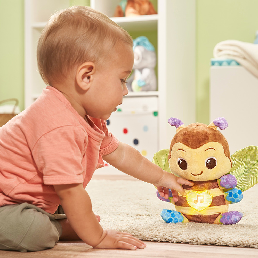 Ein Baby greift nach einem Plüsch-Bienen-Spielzeug mit leuchtendem Herzen, das auf einem Teppich sitzt.